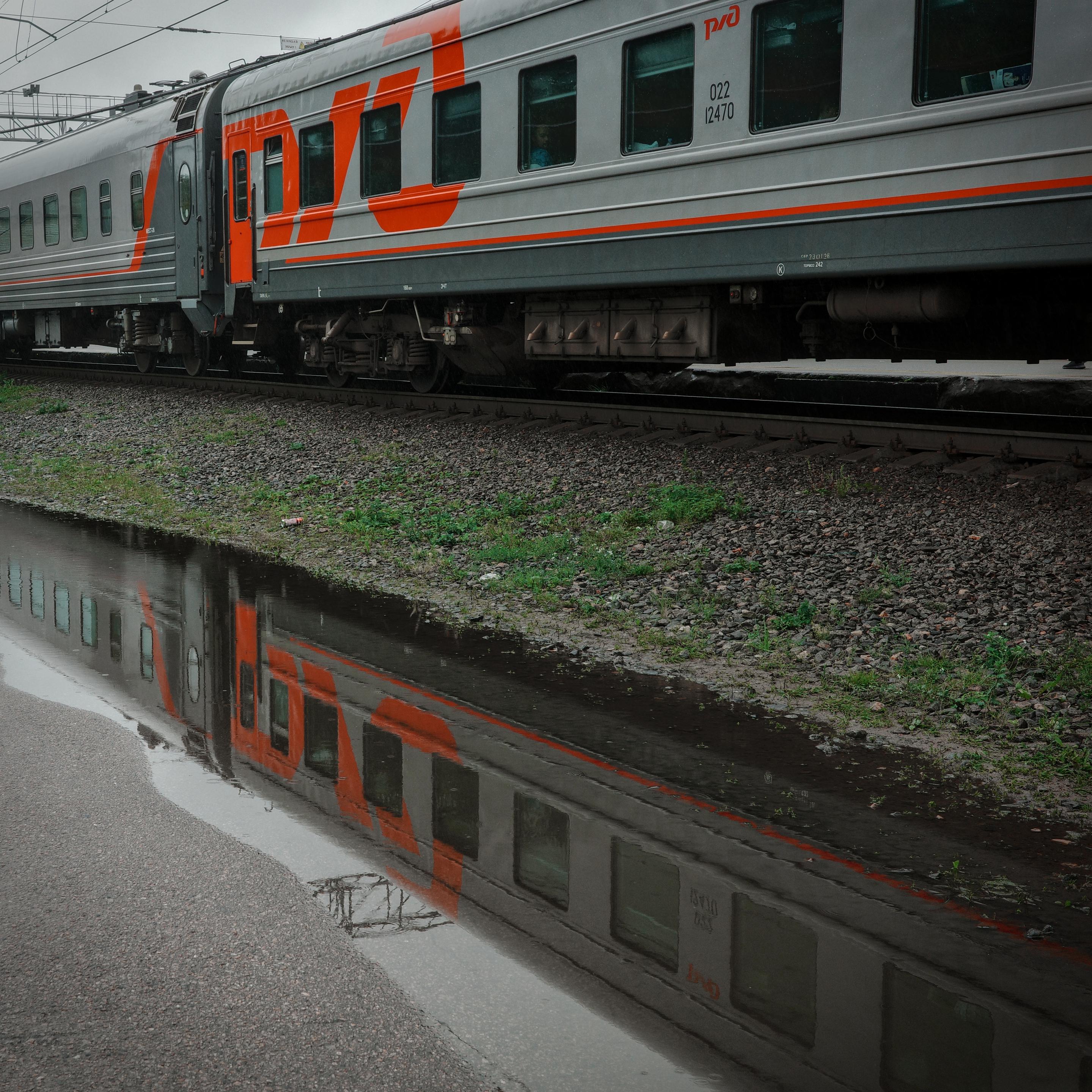An RZD train reflected in a puddle on the roadside. The dark water creates an "upside-down world" effect, and the company logo turns into a mirrored inscription. The image conveys a sense of dampness and a quiet, melancholic atmosphere.