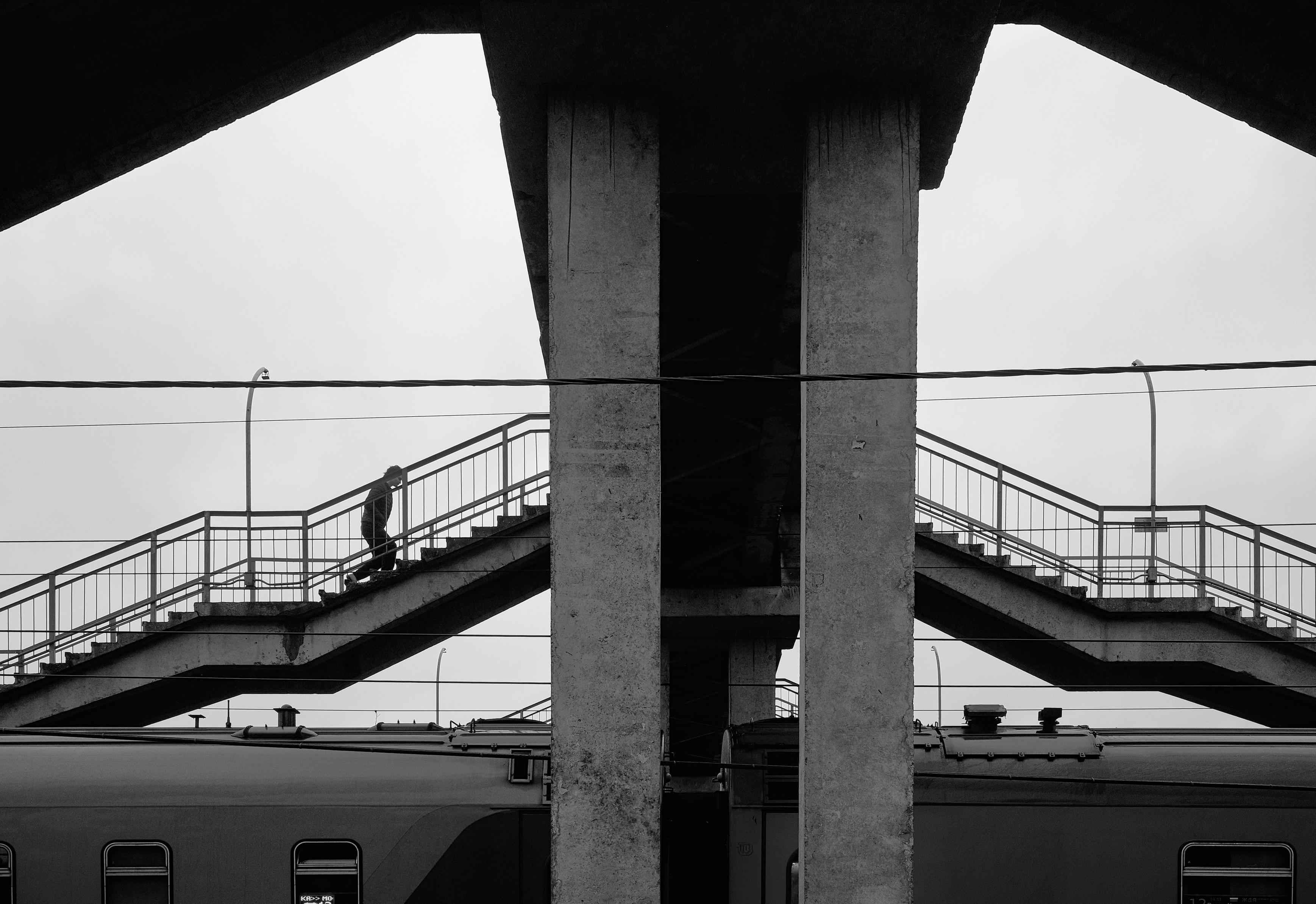 A stark, almost brutalist architecture of a concrete overpass above the tracks. Two symmetrical staircases, massive columns, and a slender human figure on one of them. Wires crossing the sky add graphic quality to the frame. The image conveys a sense of monumentality and solitude.