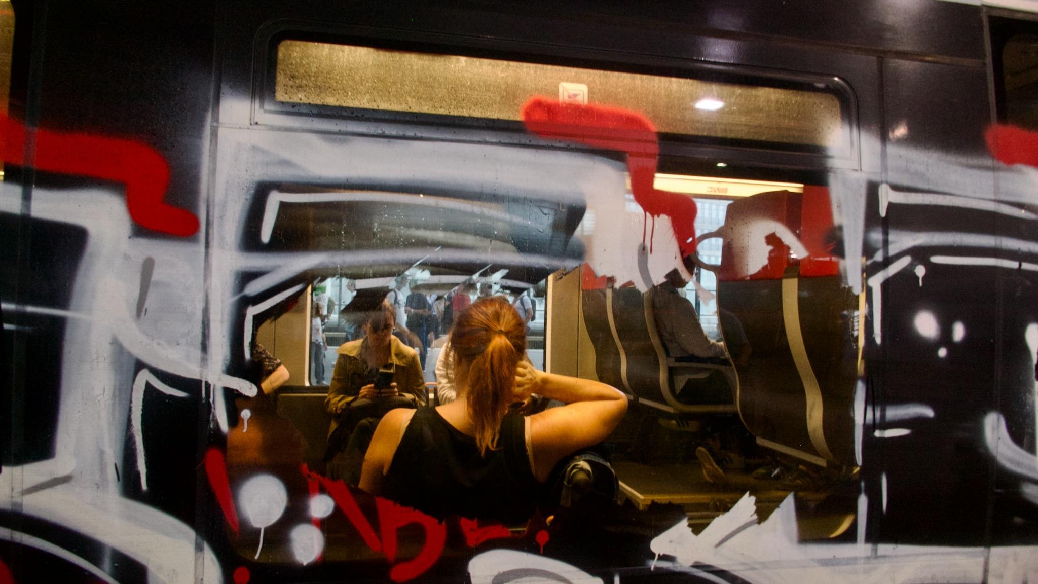 Picture shows a train wagon for persons from the outside. The big window is painted with a graffiti. In the middle the paint was scratched off to give sight from the inside. Inside the wagon is light falling on a woman sitting with the back to the window. She has her right arm raised to the head, sitting on a bench. Another woman is on the opposite side but in the background.