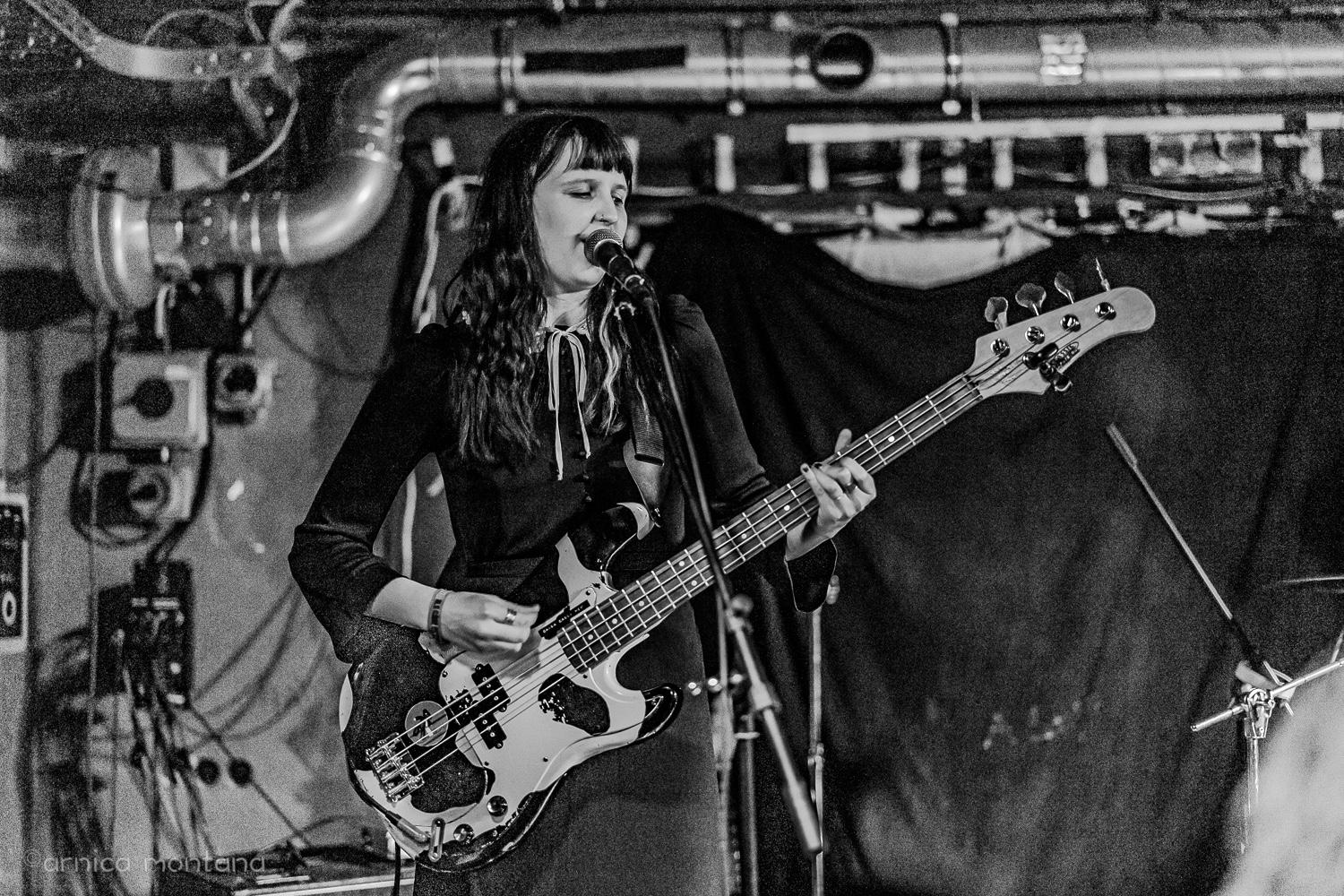An artist is performing on a stage with chaotic cables, switches and pipes visible in the background on a black and white picture. She is singing into a microphone and playing bass guitar.