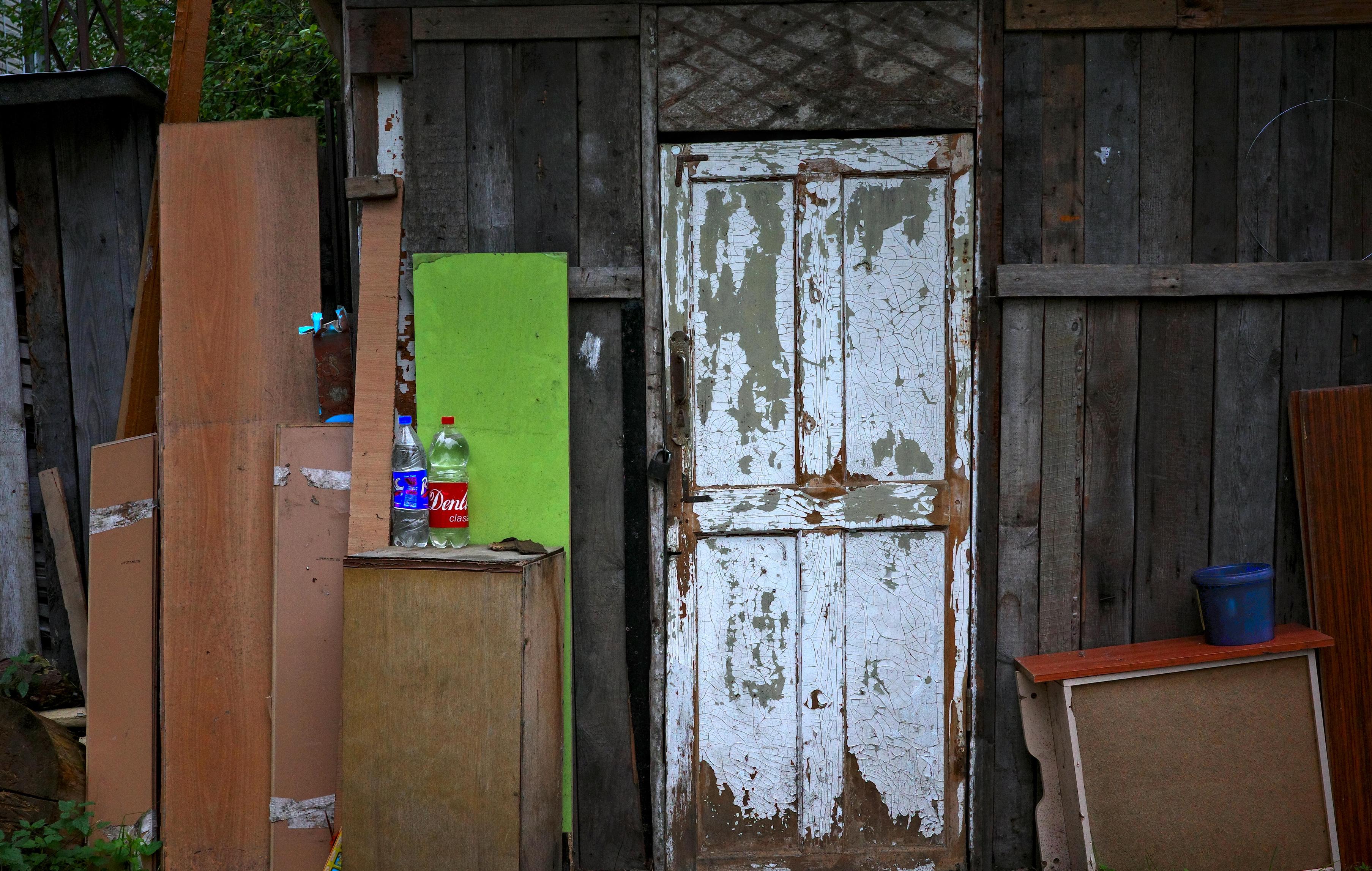 This color photograph captures the wall of an old utility building or shed, which has turned into a veritable visual collage of heterogeneous objects and materials. In the center of the composition is an old wooden door painted white, with cracked and peeling paint revealing dark wood underneath. To the left of the door, on a wooden panel, stand two plastic bottles — one with water and one with "Dyushes" soda. Next to them, a bright green sheet of plywood is propped up, creating a sharp color accent. Around it is a jumble of wooden planks, panels, old furniture, and a blue plastic bucket. This entire heterogeneous "mosaic" of textures and colors forms a single composition that can be studied endlessly. The image conveys the atmosphere of a cluttered but cozy and very "human" everyday life.