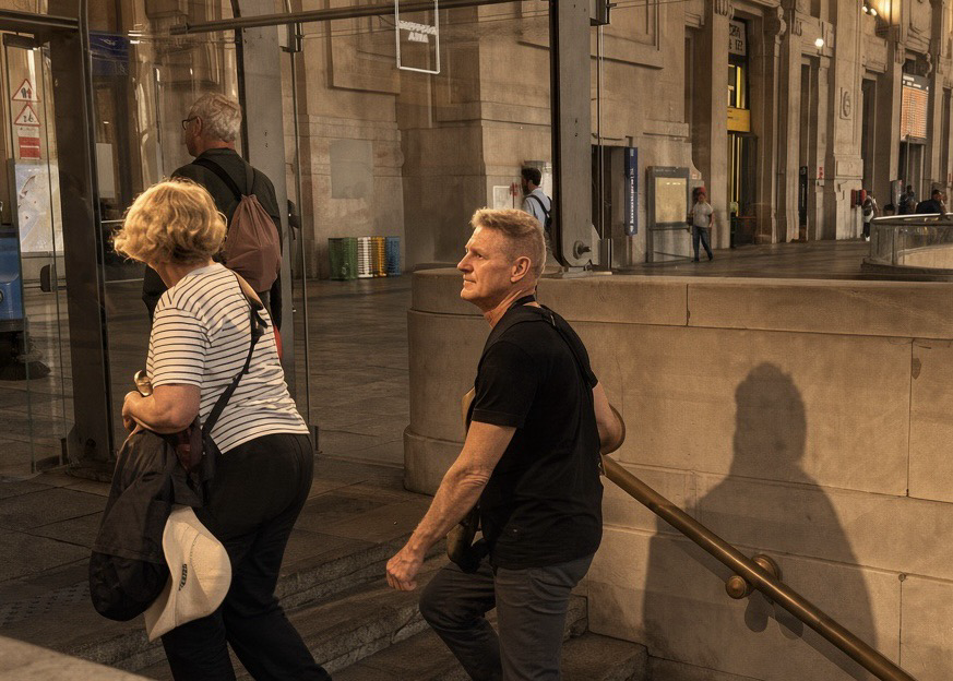Candid street photo of commuters on stone stairs in a covered European station hall. A woman in a striped shirt carries a backpack and a large bag; a man in black looks back over his shoulder; another man walks ahead. Warm light, glass walls, and a long shadow on the wall.