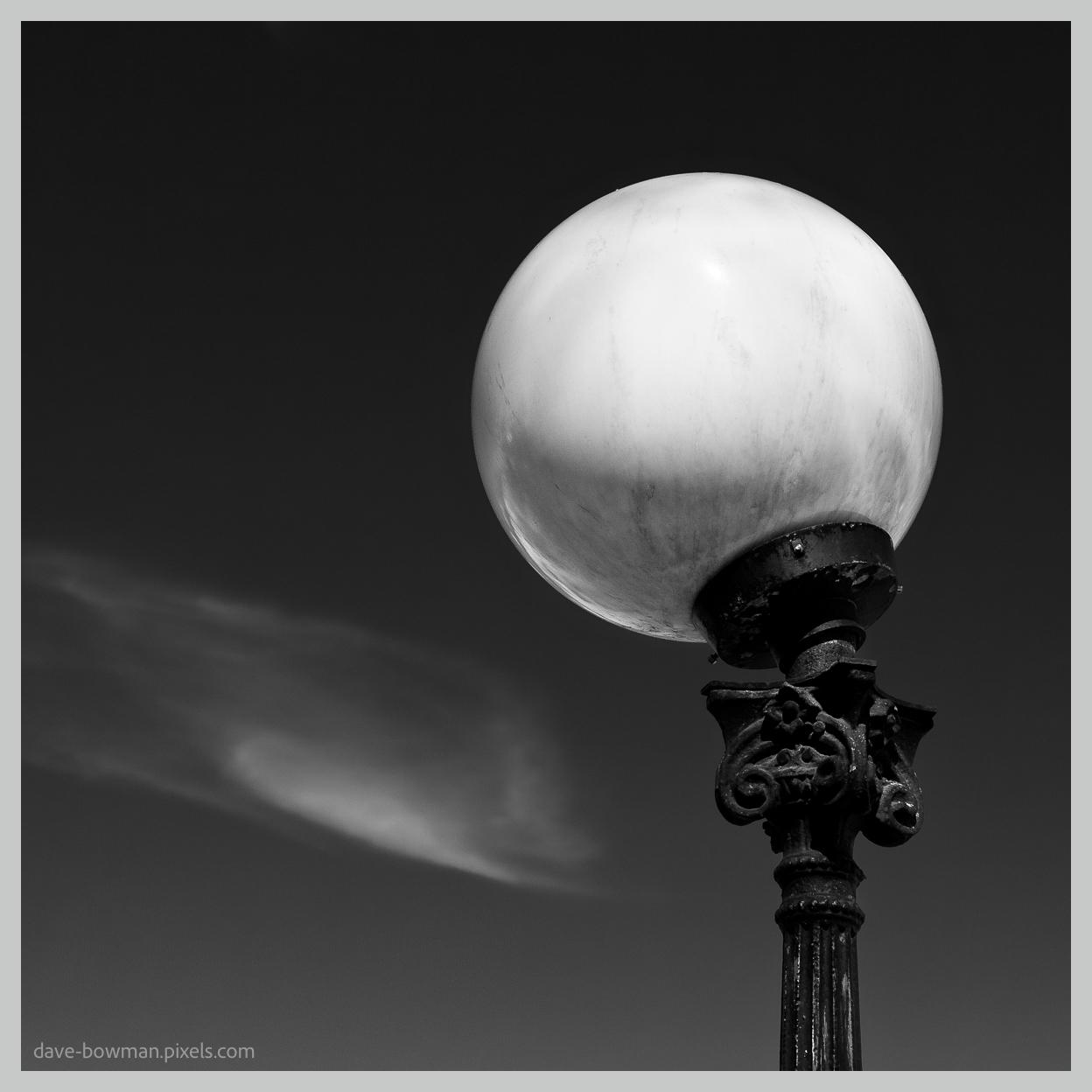 A monochrome photograph of a white globe-shaped street lamp standing against a dark Stockholm sky, with its smooth, rounded form contrasting sharply with the ornate dark metal structure supporting it. A single streak of cloud passes across the sky behind the lamp, adding a subtle sense of movement to the otherwise simple composition.