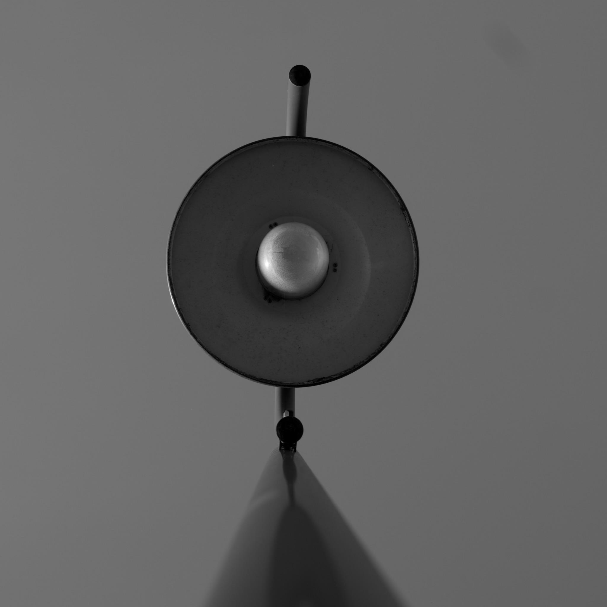Picture of a street light lantern from the ground straight up. In the middle of the photo is the light bulb, surrounded by a metal sheet in a circle. On the bottom of the picture is the pole. Background is plain blue sky.
Photo is in black and white.