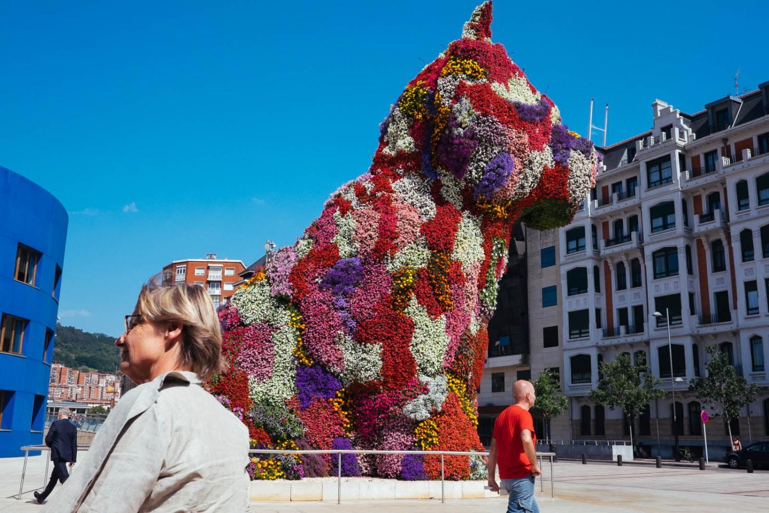 Photograph of Jeff Koons' *Puppy* next to the Guggenheim Museum Bilbao, with several people walking past.