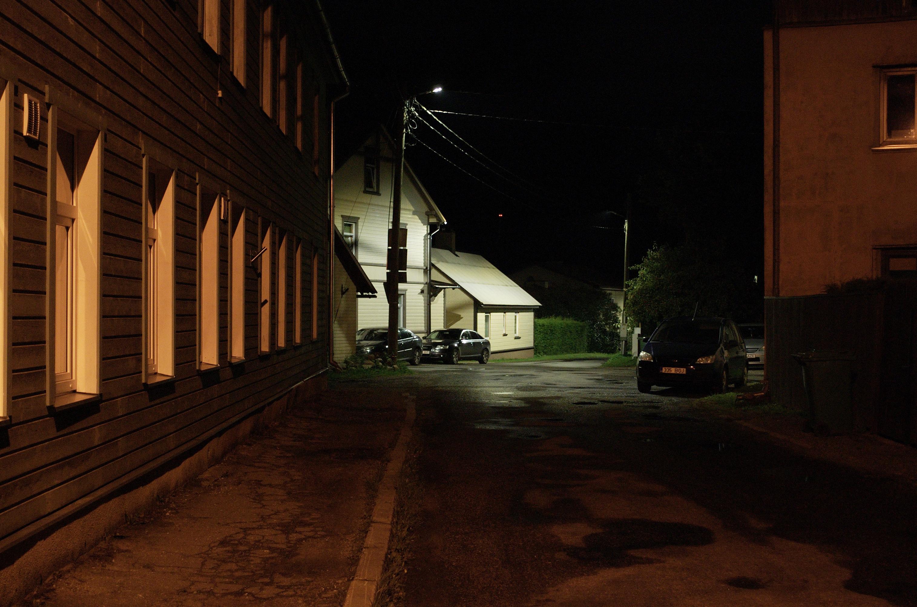 Photo of a street at night. To the left runs the grey wall of a two-storey wooden house. The sodium streetlight behind me casts a dim orange glow on it, highlighting the white windowframes. The rest of the frame is mostly dark, but a newer white LED light down the street picks out two beige houses in sharp contrast.