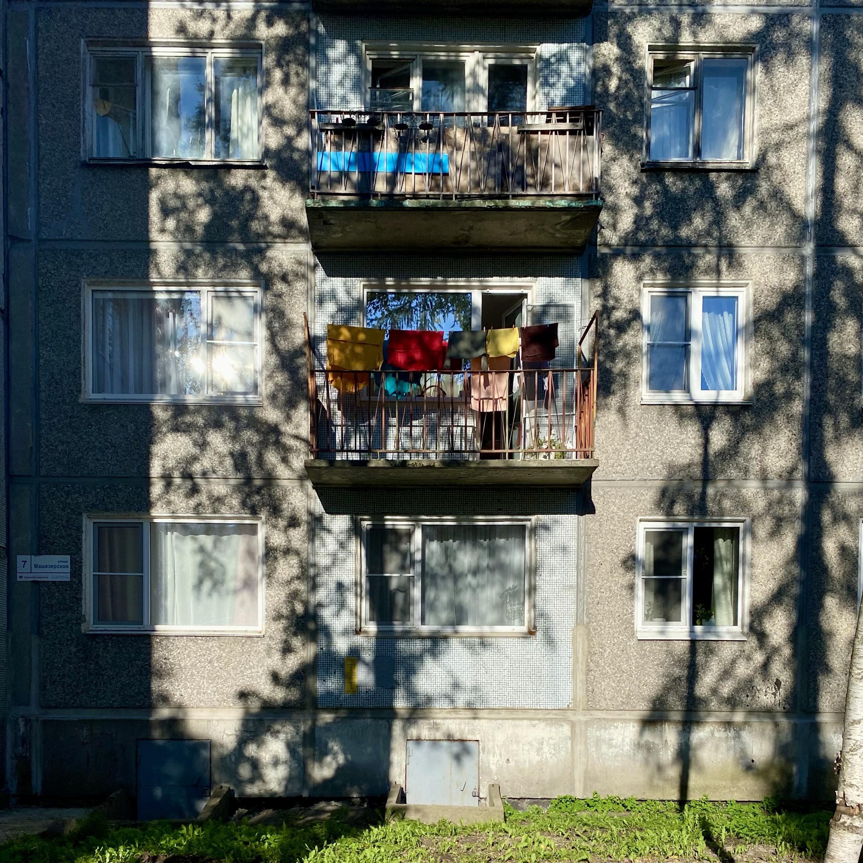We see the façade of an ordinary city apartment block, flooded with bright sunlight. What gives the shot its particular mood are the deep shadows of tree branches. They weave a whimsical, living pattern that slides across the walls and windowpanes.

At the heart of the composition is a balcony. Colourful laundry hangs there to dry — vivid yellow, red, and brown patches of fabric. This detail lends the frame warmth and a feeling of everyday life. Down below, right at the foot of the building, lush green grass grows.