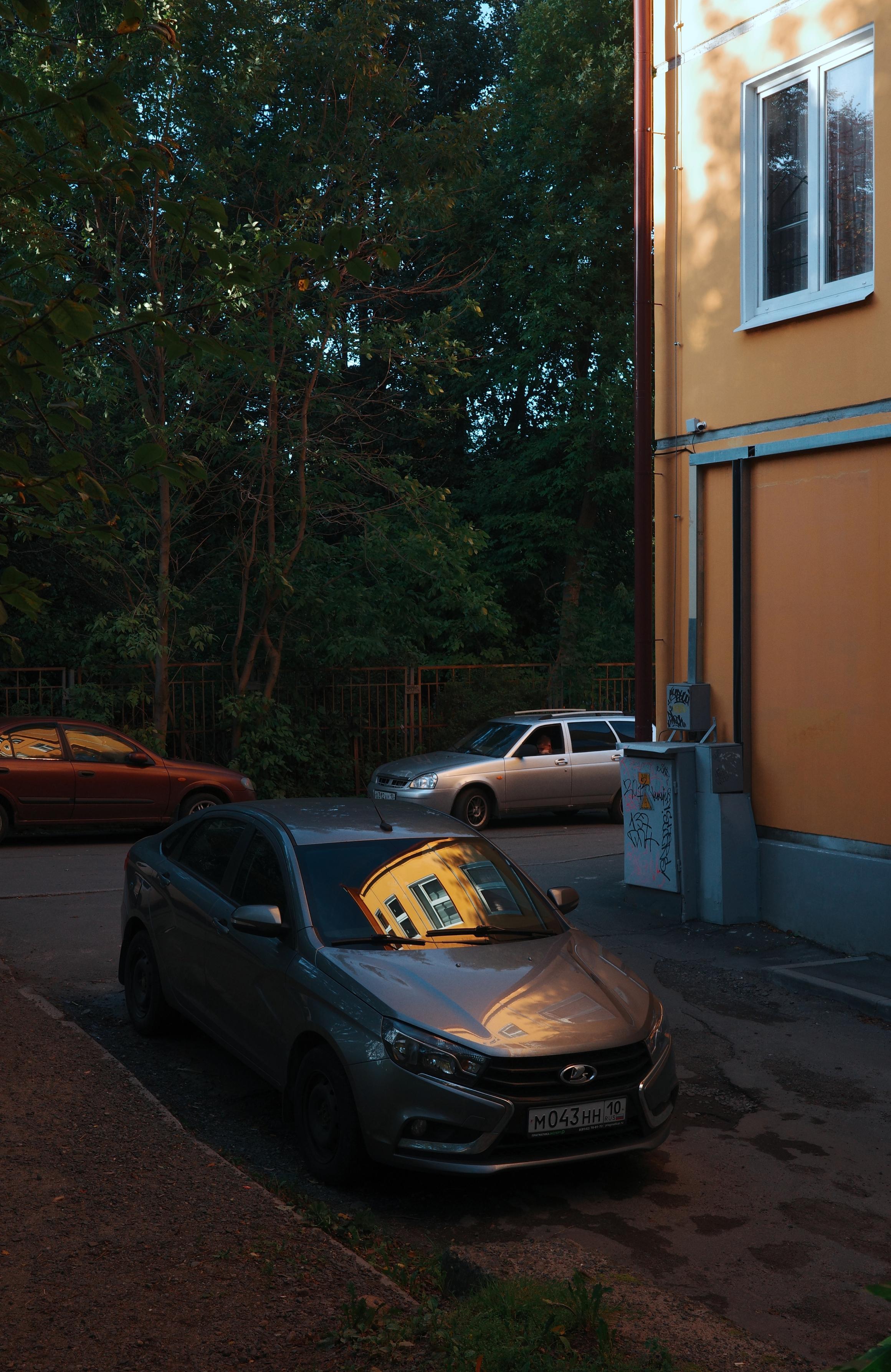This color photograph captures an evening courtyard in a quiet urban neighborhood. In the center of the composition is a gray car parked at the edge of the road. The main detail of the shot is the bright, golden reflection of a neighboring yellow house in the car's windshield, resembling a tiny sunset caught in the glass. On the right rises the facade of that same yellow building with a white window, with the warm light of the sunset falling on its wall. In the background, other parked cars are visible — a silver one and a dark red one — along with dense green trees and a fence. A gray electrical box with graffiti stands by the wall of the house. The image conveys the atmosphere of a quiet, warm evening and the coziness of an urban courtyard, where the light of sunset transforms ordinary things.