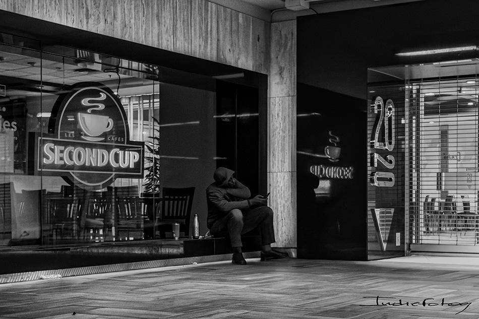 A man is sitting on the ground, beside a closed coffee shop with his own coffee at night.