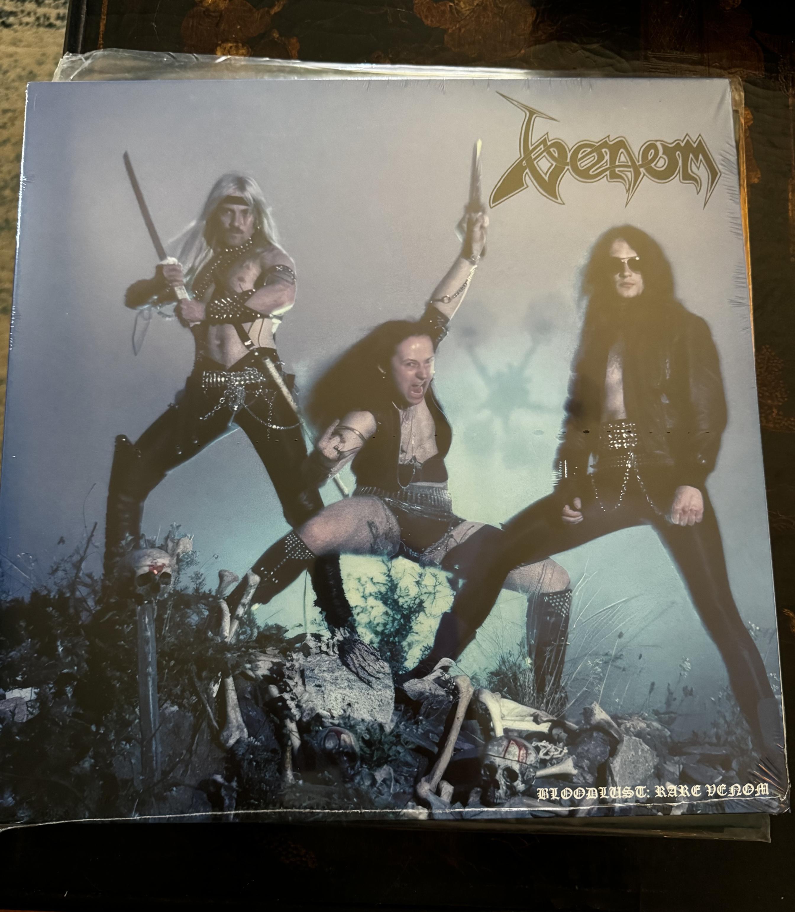 Front cover of bootleg album Bloodlust: Rare Venom.  The band trio stand atop rocks and broken bones. Mantas wields a sword and leather pants, Cronos throws a shape in the middle, and Abaddon stands with his aviator sunglasses on. 