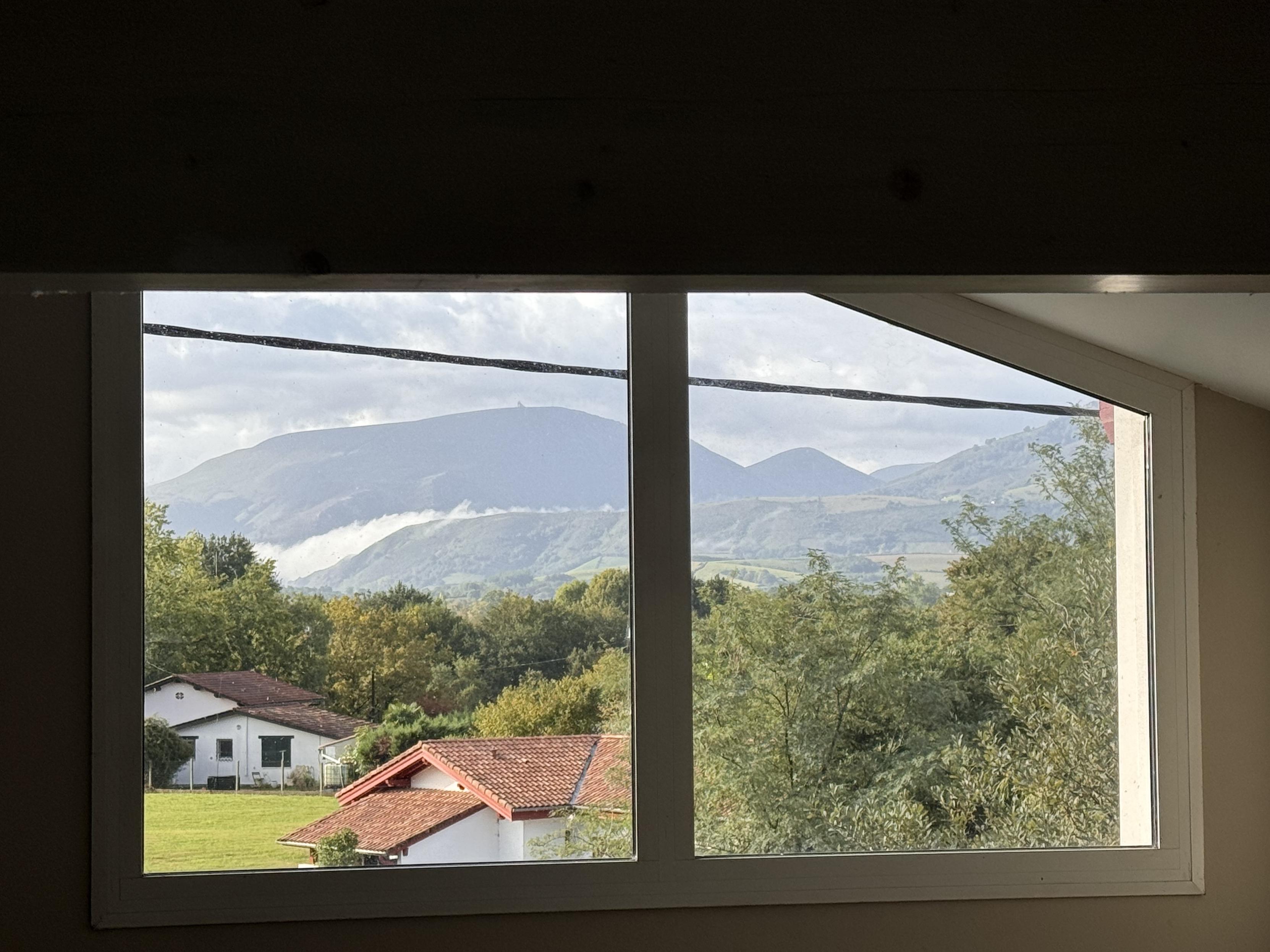 A view from a window showing rolling mountains with a layer of white clouds between them, and residential houses with red roofs in a green landscape, under a partly cloudy sky but with sun light coming from the left.