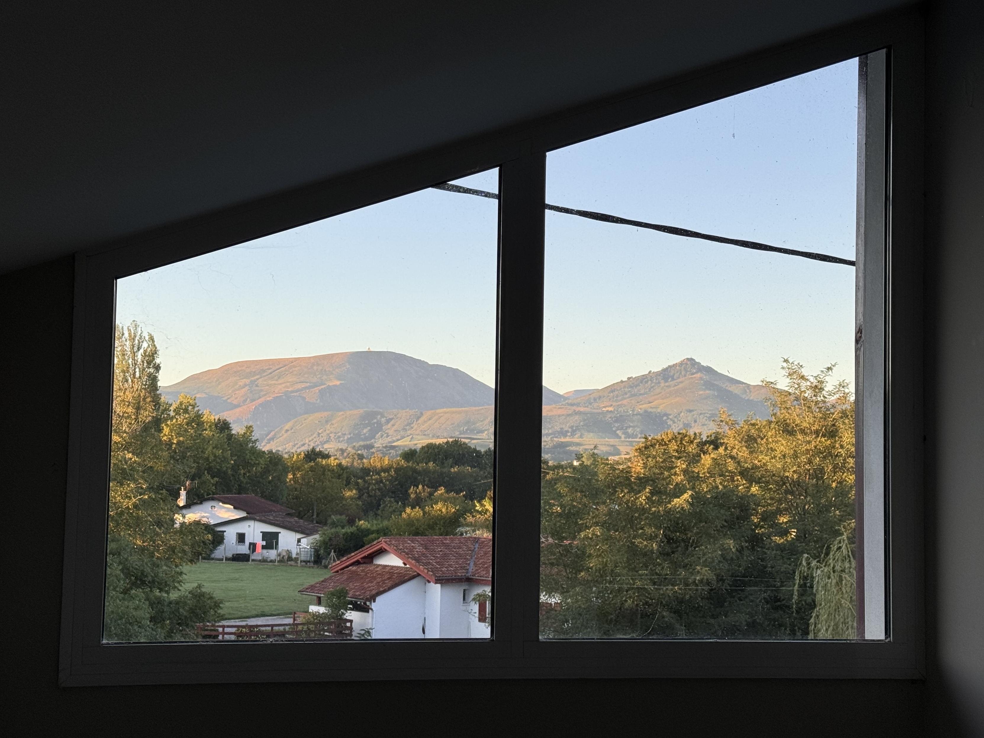 A view from a window showing rolling mountains in the background and residential houses with red roofs in a green landscape, under a clear blue sky as the morning sun is casting warm light on trees and the mountains.