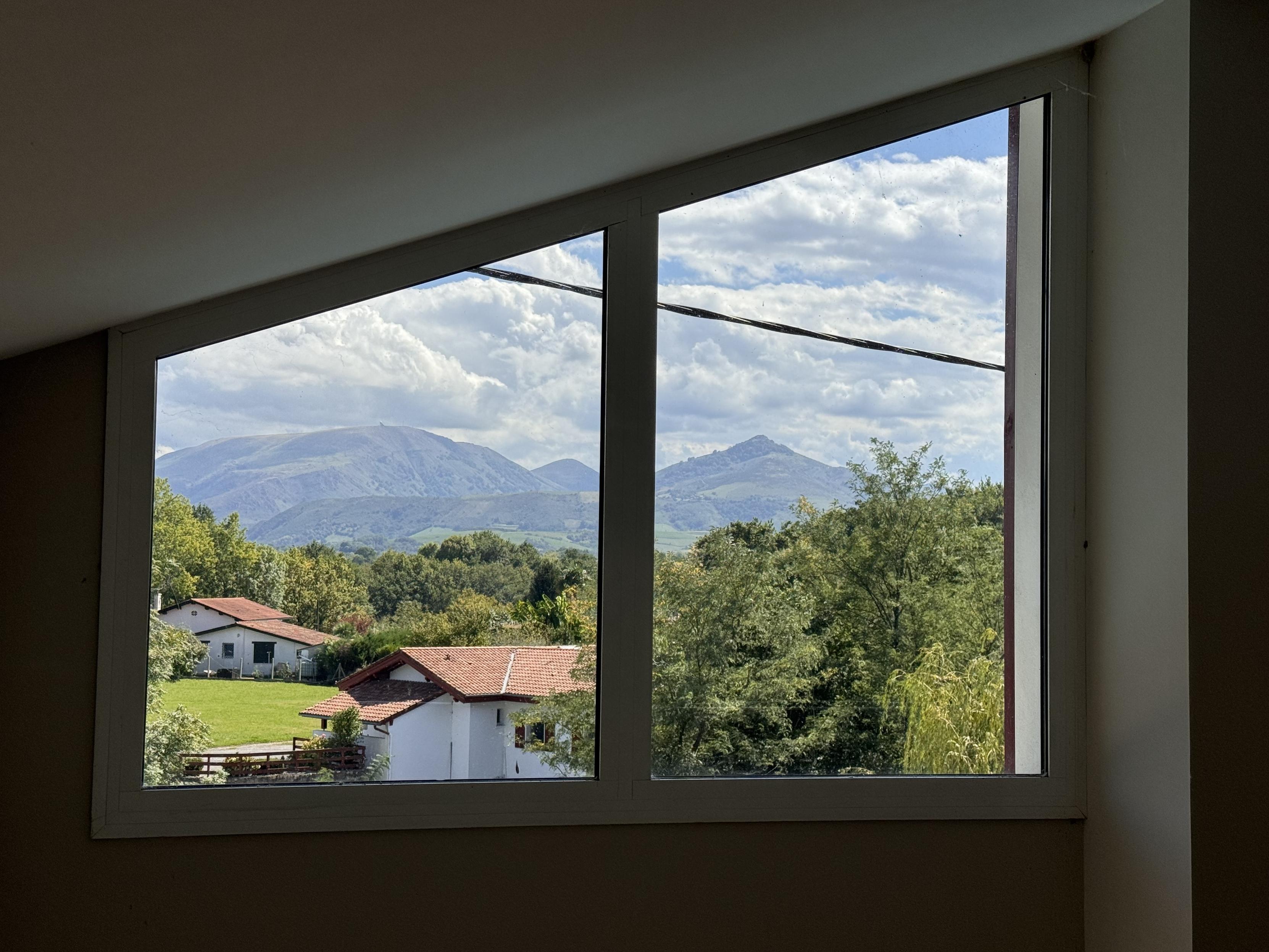 A view from a window showing rolling mountains in the background and residential houses with red roofs in a green landscape, under a partly cloudy sky.