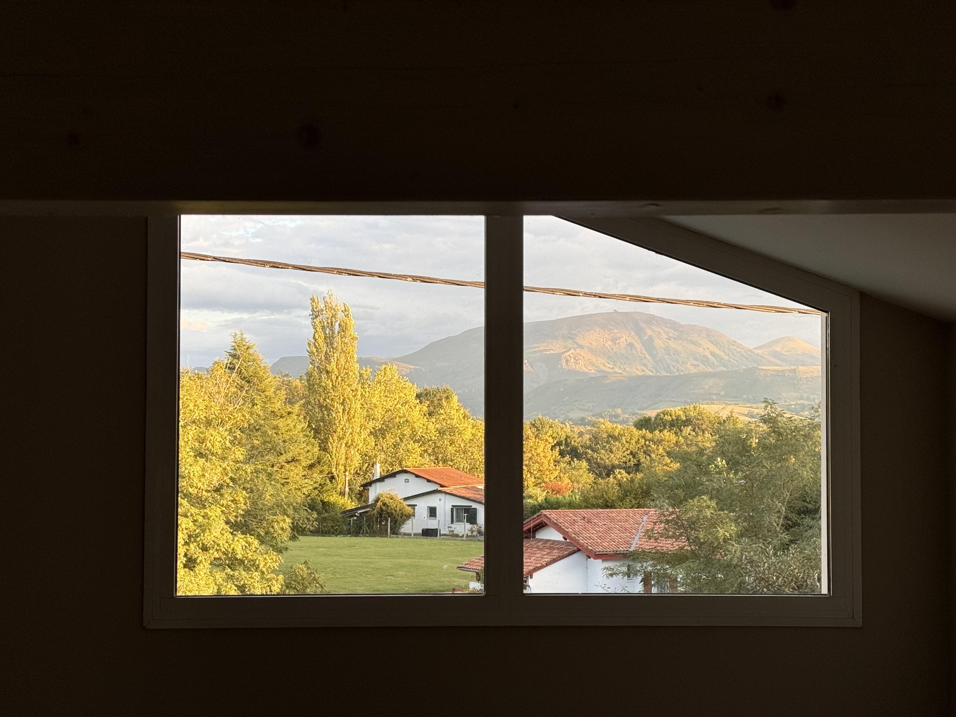 A view from a window showing rolling mountains in the background and residential houses with red roofs in a green landscape, as the setting sun is casting a warm light on the trees and the mountains.