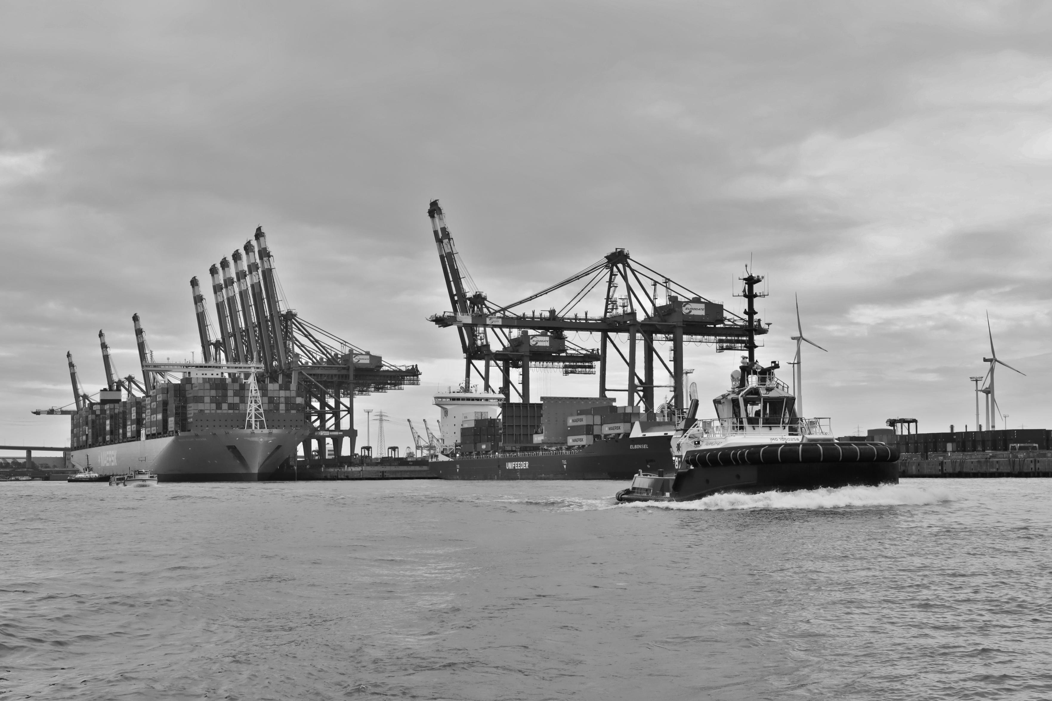 Schwarzweiss-Foto einer typischen Hafenszene in einem Containerhafen in Hamburg mit Containerschiffen und Be- und Entladebrücken im Hintergrund (links) sowie einem vorbeifahrenden Hafenschleppers im Vordergrund (rechts).
