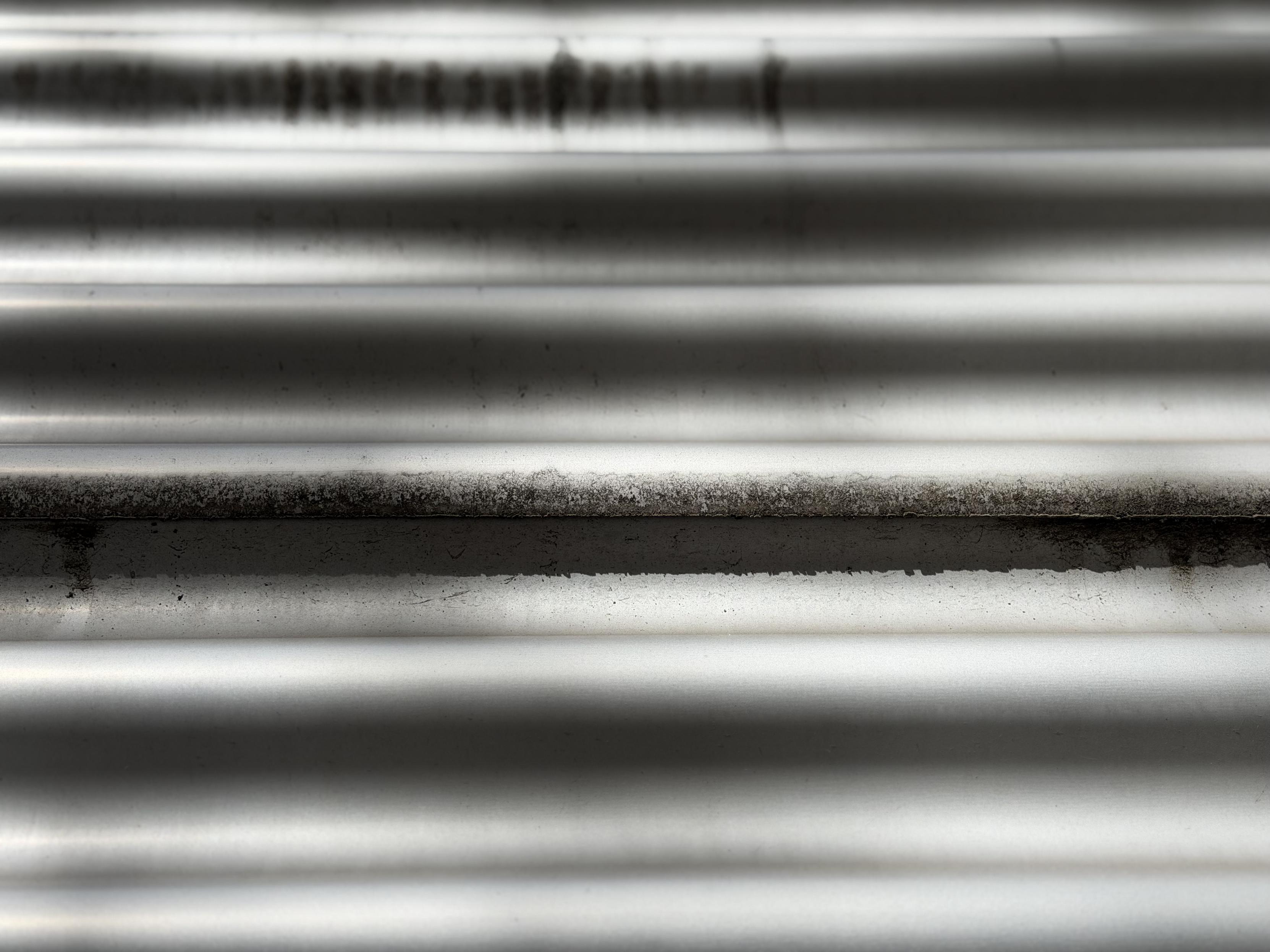 Close-up of a horizontally corrugated metal surface, likely corrugated sheet metal, in light grey to silver tones. The ridges run parallel across the entire frame, catching raking light in alternating bright and dark stripes. Along one of the upper ridges, a blurred dark pattern is visible that resembles lettering or printing. In several places, especially toward the middle of the image, dark, crumbly grime or deposits cling to the raised edges.