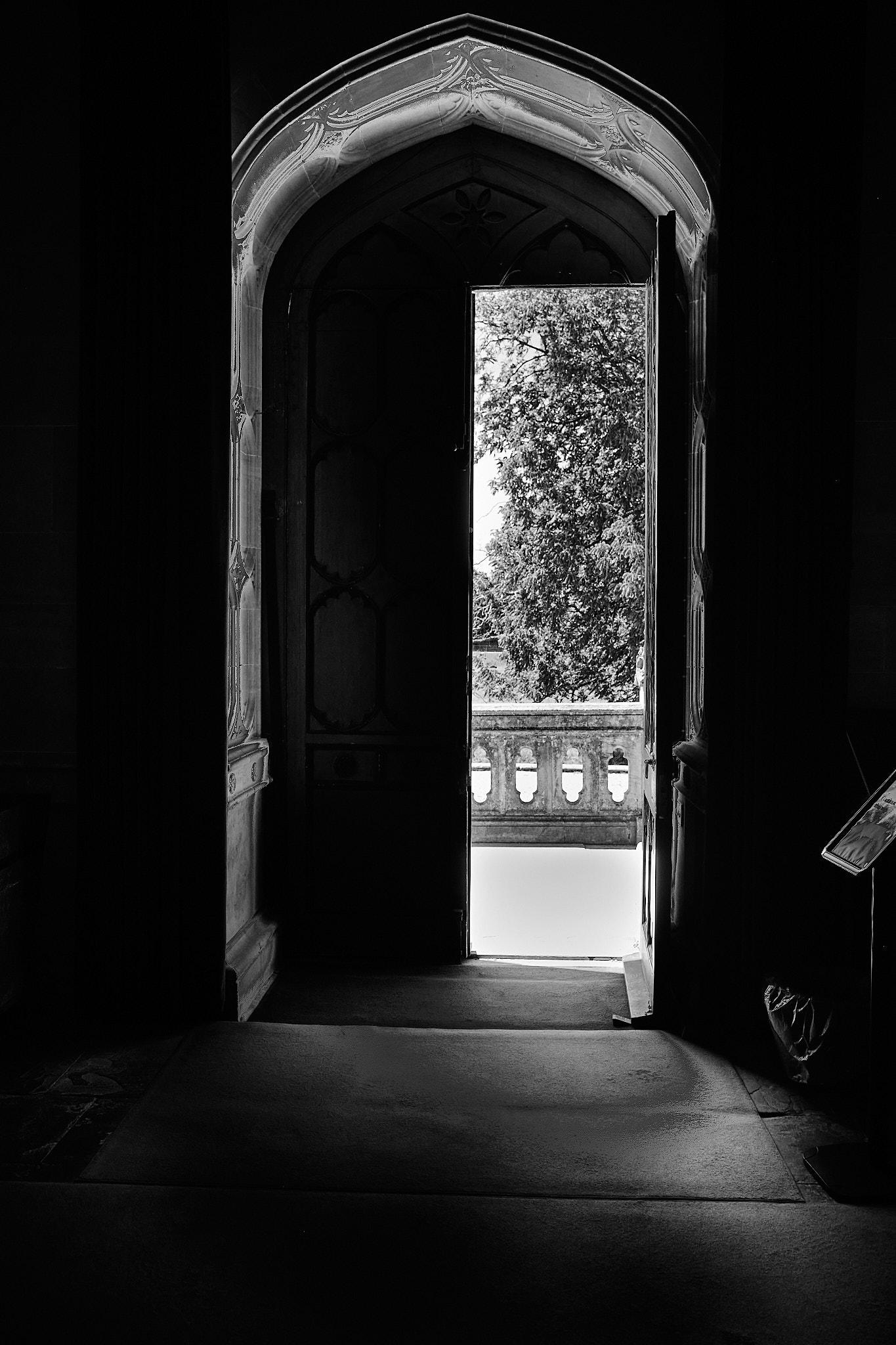 An open door is visible in the foreground, framed by ornate architecture. Bright sunlight streams through the doorway, illuminating the dark interior. Outside the door, a tree and a balcony railing are partially visible.