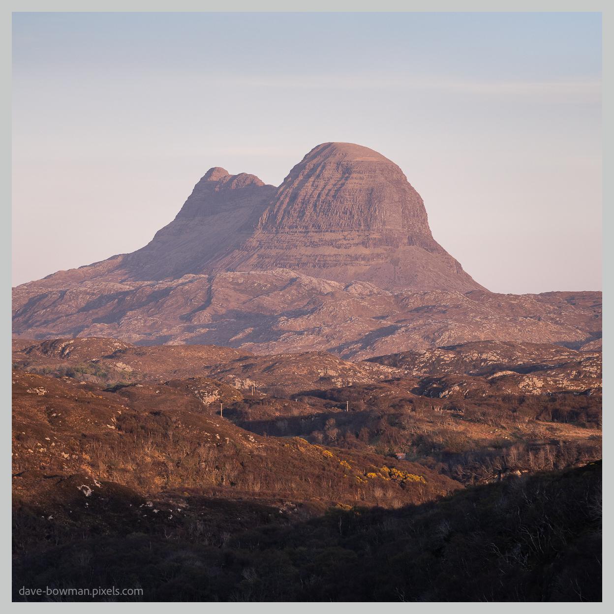 A photograph of Suilven in Assynt, in the Highlands of Scotland, rising prominently above the surrounding open moorland. The distinctive mountain has a rugged sandstone profile with a series of steep, rounded peaks, highlighted by warm evening light that gives its rocky slopes a soft golden glow. Standing isolated from the surrounding hills, Suilven's unusual shape dominates the landscape and creates a strong focal point against the open terrain.