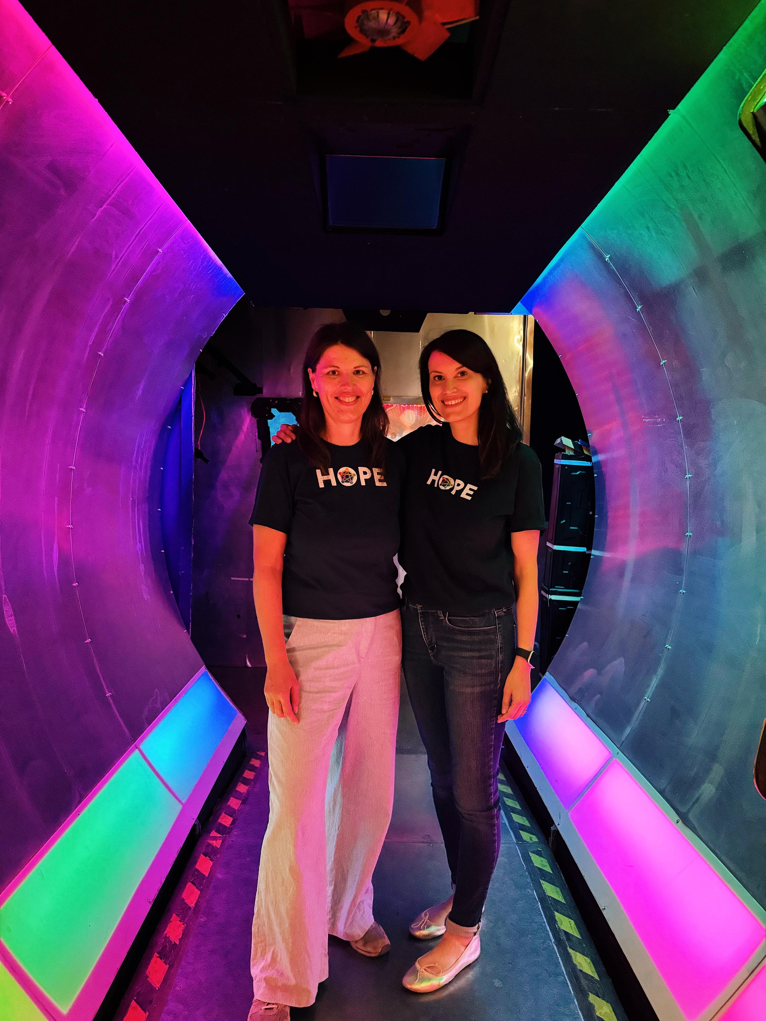A photo taken at C-base’s iconic entrance in a tunnel with colorful neon lights (magenta, green and blue tint). On the left you see Samvie wearing a blue t-shirt with the word HOPE and the Fediverse logo inside the O… and next to her yours truly with the same HOPE t-shirt
