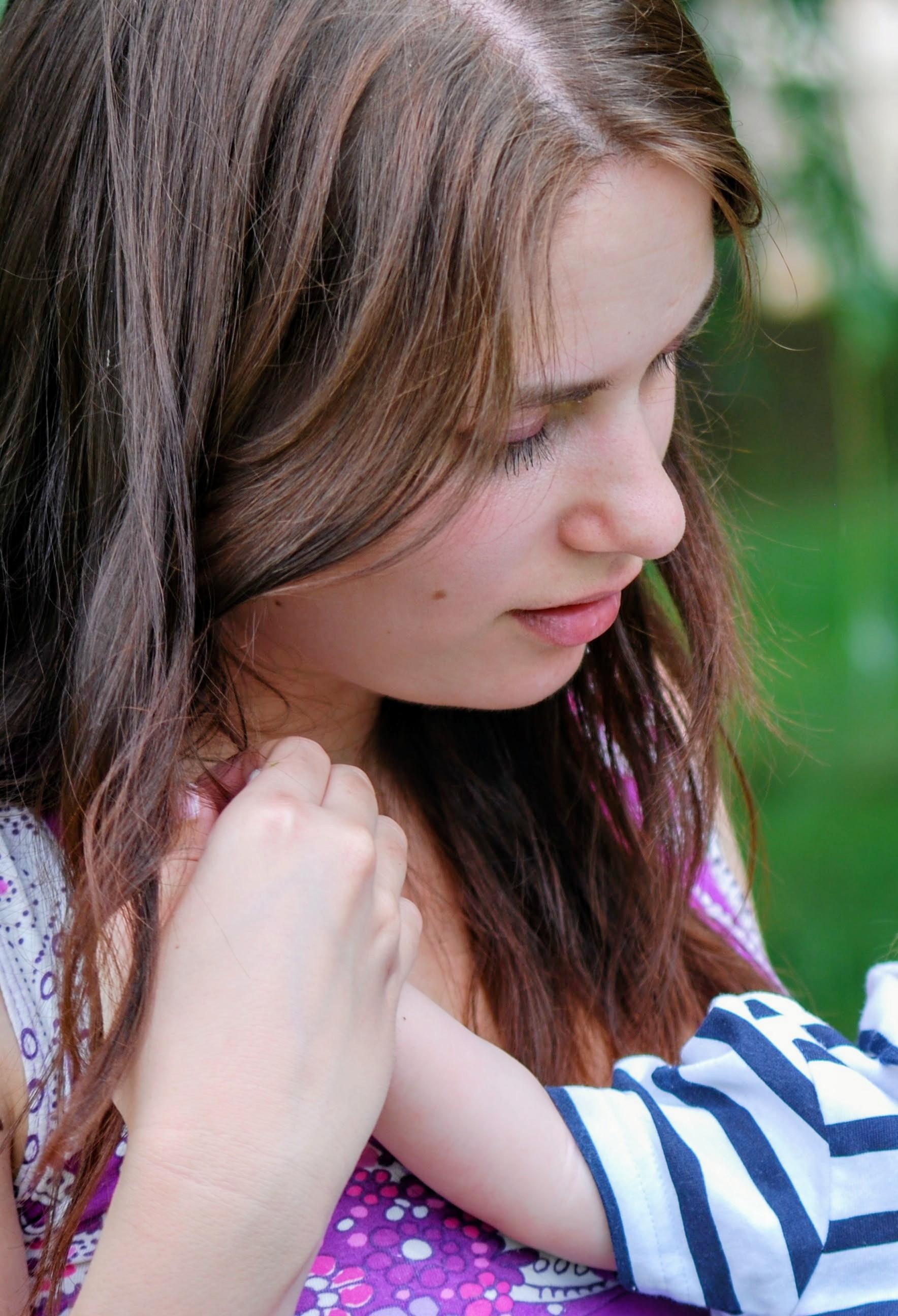 A close-up portrait shows a young woman with brown hair looking down, holding her hand near her collarbone while wearing a purple and white patterned top. A child wearing a blue-and-white striped sleeve reaches toward her.