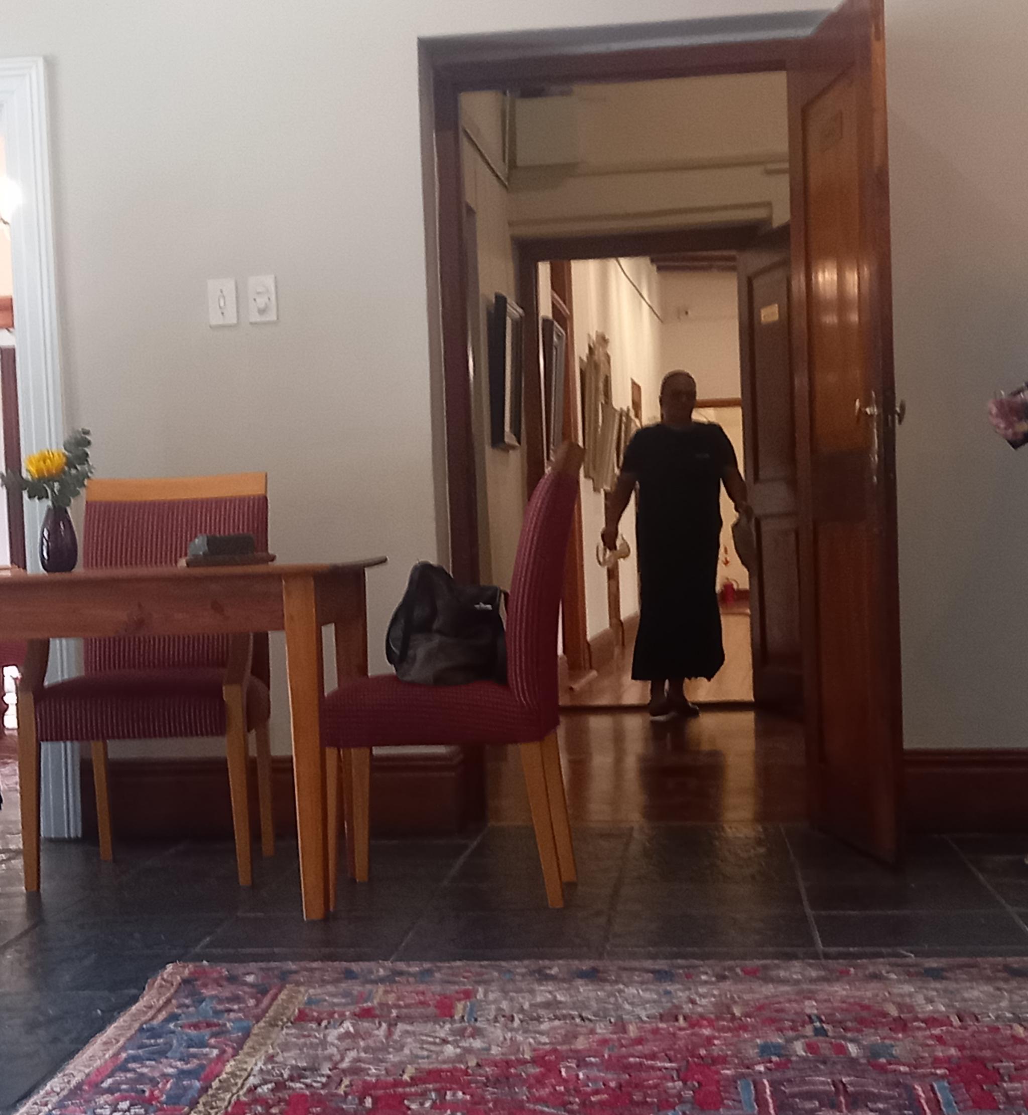 A woman in a long dress is seen through a door  walking down a long passageway 
The floors are highly polished and a carpet covers part of the floor
There is a desk and chair on the left of the image