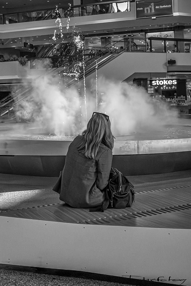 A woman is sitting in front of a fountain with a foggy mist blocking  her view.