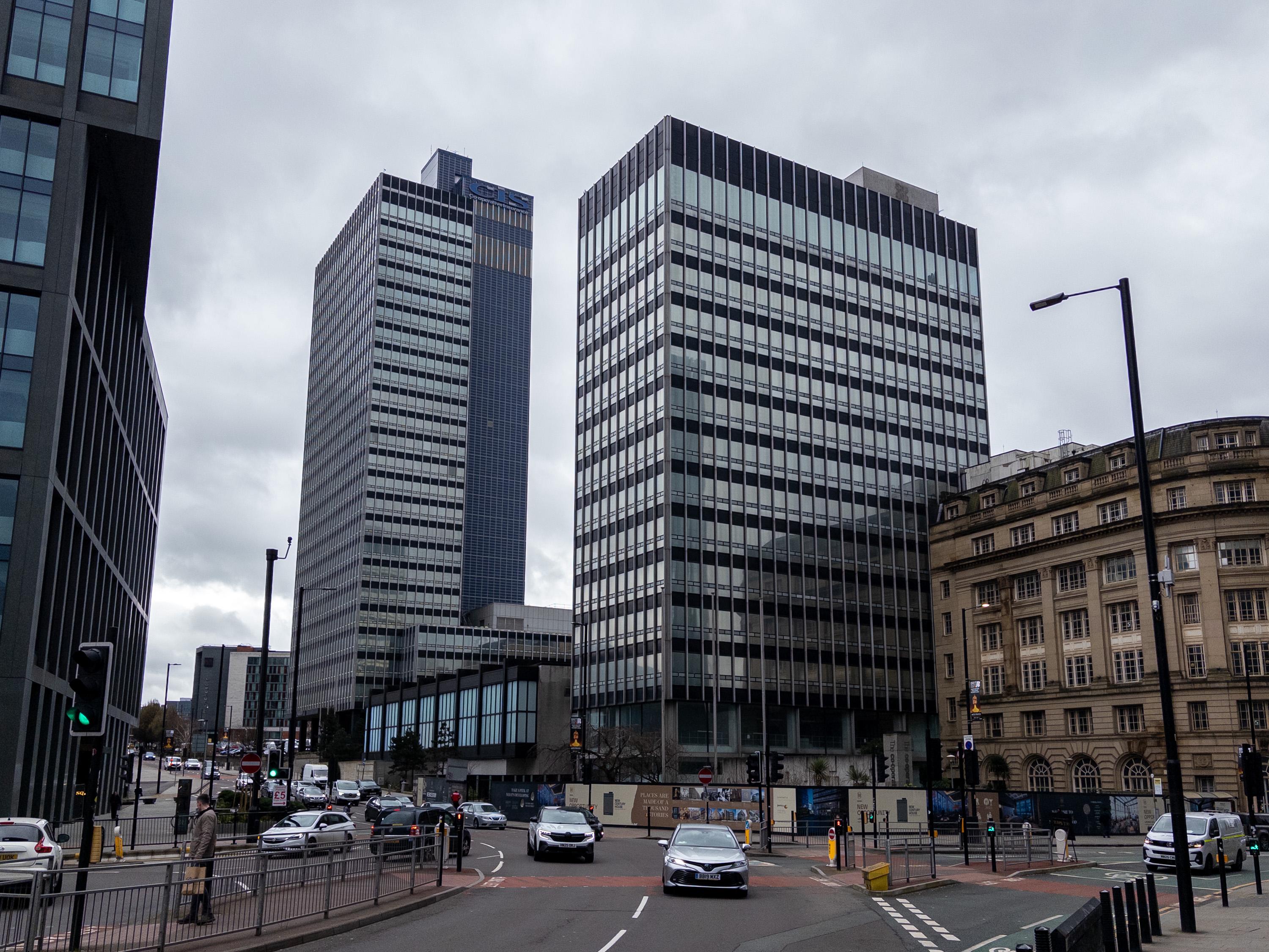 A city street with cars driving and tall, modern office buildings with glass windows under a cloudy sky. To the right, an older stone building with a curved façade is visible. Pedestrians walk on pavements and zebra crossings in the foreground.