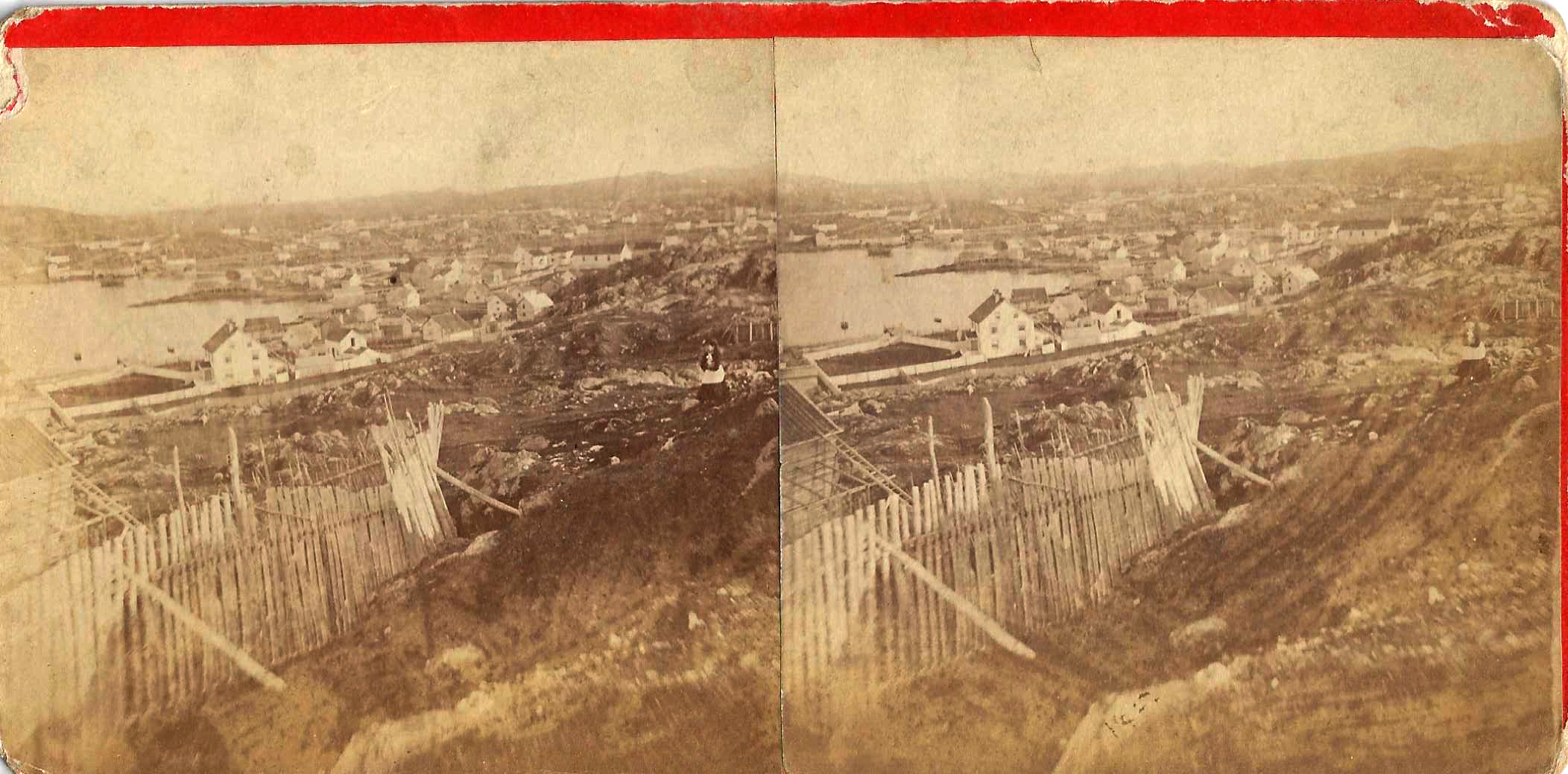 A side-by-side photo of Twillingate, Newfoundland from a card intended to be viewed in a stereoscope. The photo is very faded and shows the harbour on the left, with the houses surrounding it. In the foreground is a rickety fence and (barely visible) is a woman standing the rocky field.