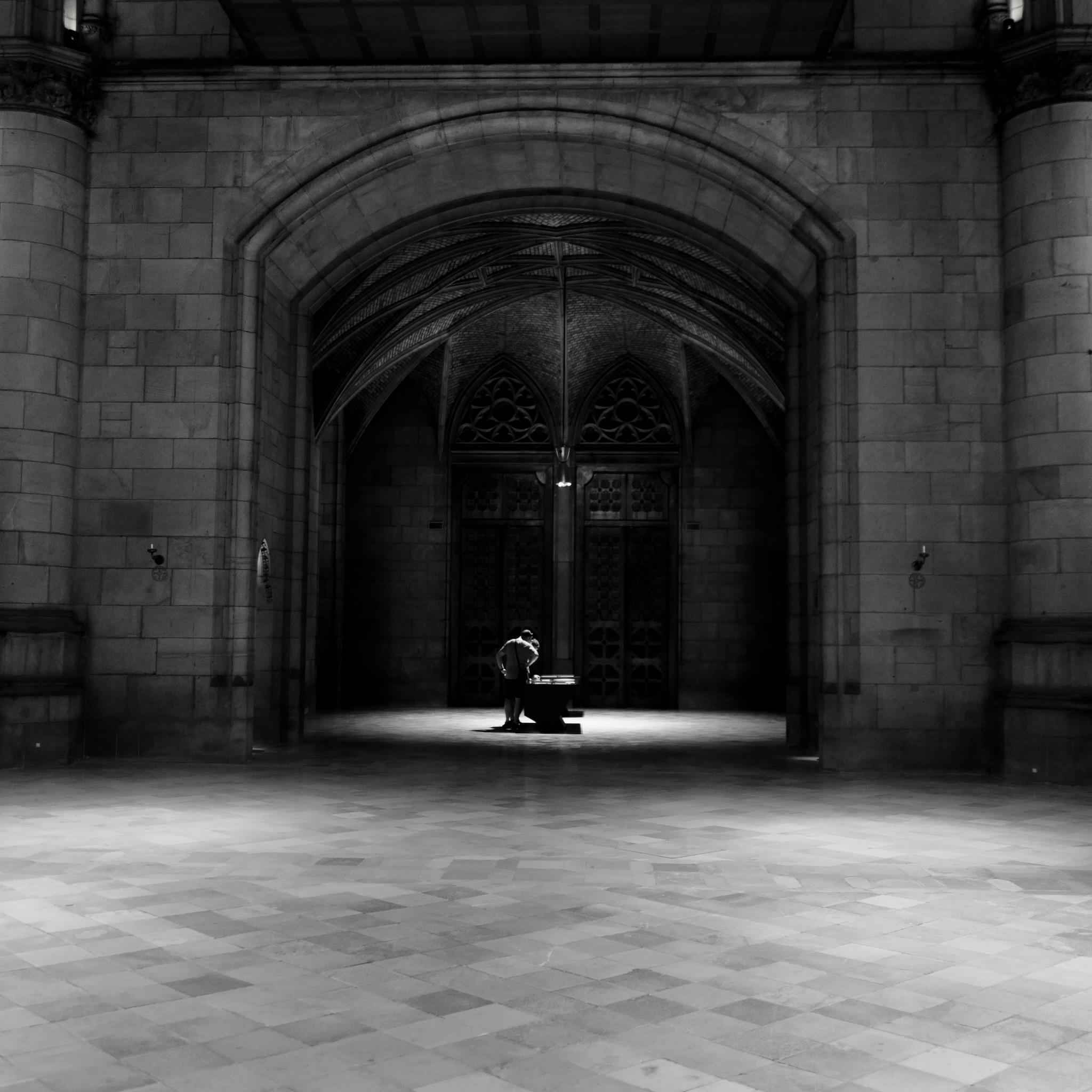 Picture taken in the New Cathedral in Linz, Austra. In the center is a table, illuminated by a spotlight from above. A person is standing beside the table on the left. The room is operated by an entrance from the main church room. The floor seems endless in all directions.
Photo is black and white.