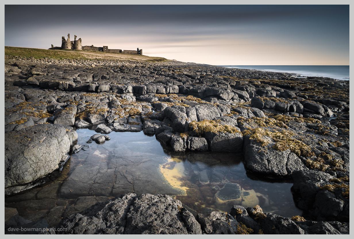 A photograph of Dunstanburgh Castle in Northumberland, England, viewed from the coast at dawn, with the historic castle standing on the horizon in the distance. A calm rock pool occupies the foreground, reflecting the soft early morning light, while the surrounding shoreline creates a quiet and peaceful coastal scene.