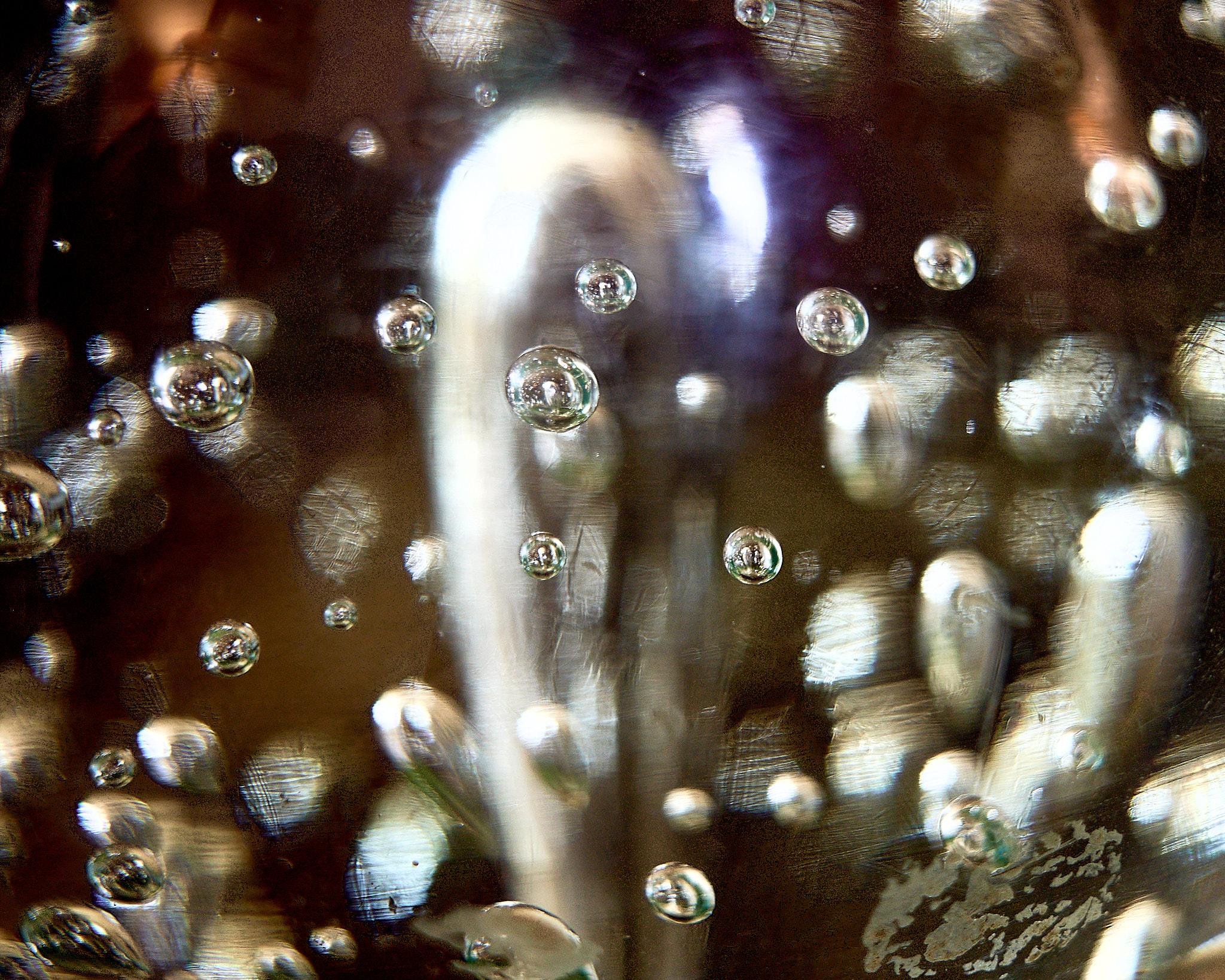 A close-up view of a glass object filled with bubbles. The glass surface is reflective, showcasing multiple spherical bubbles of varying sizes. The background appears blurred with light reflections, creating a shimmering effect.