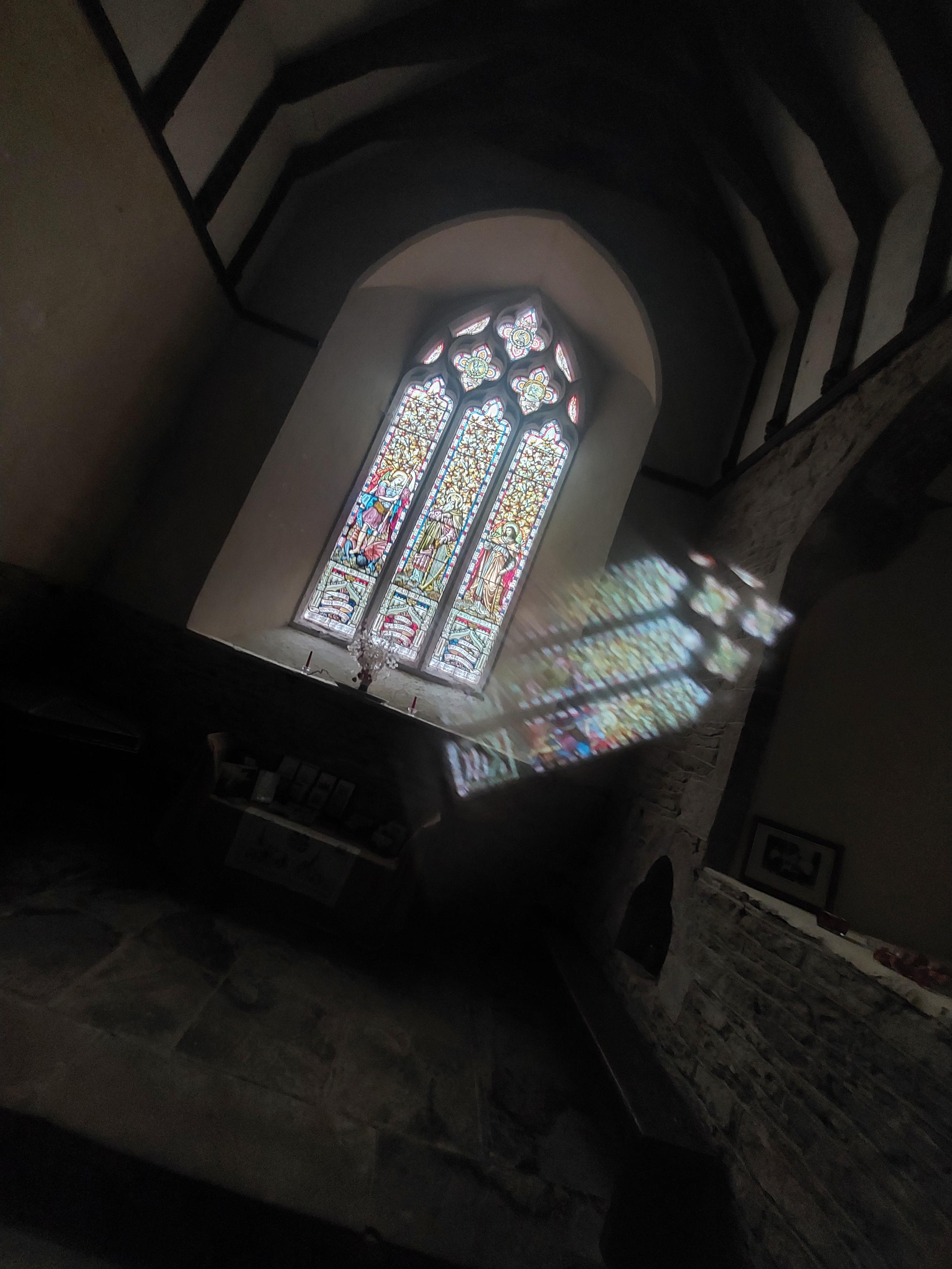 Inside a small old church, sunlight streams through a tall stained-glass window, projecting a vivid, ghostly reflection of the window across the rough stone wall. Dark roof beams and the shadowy interior frame the luminous colours; the unusual doubled effect was caused by a crack in the camera lens.