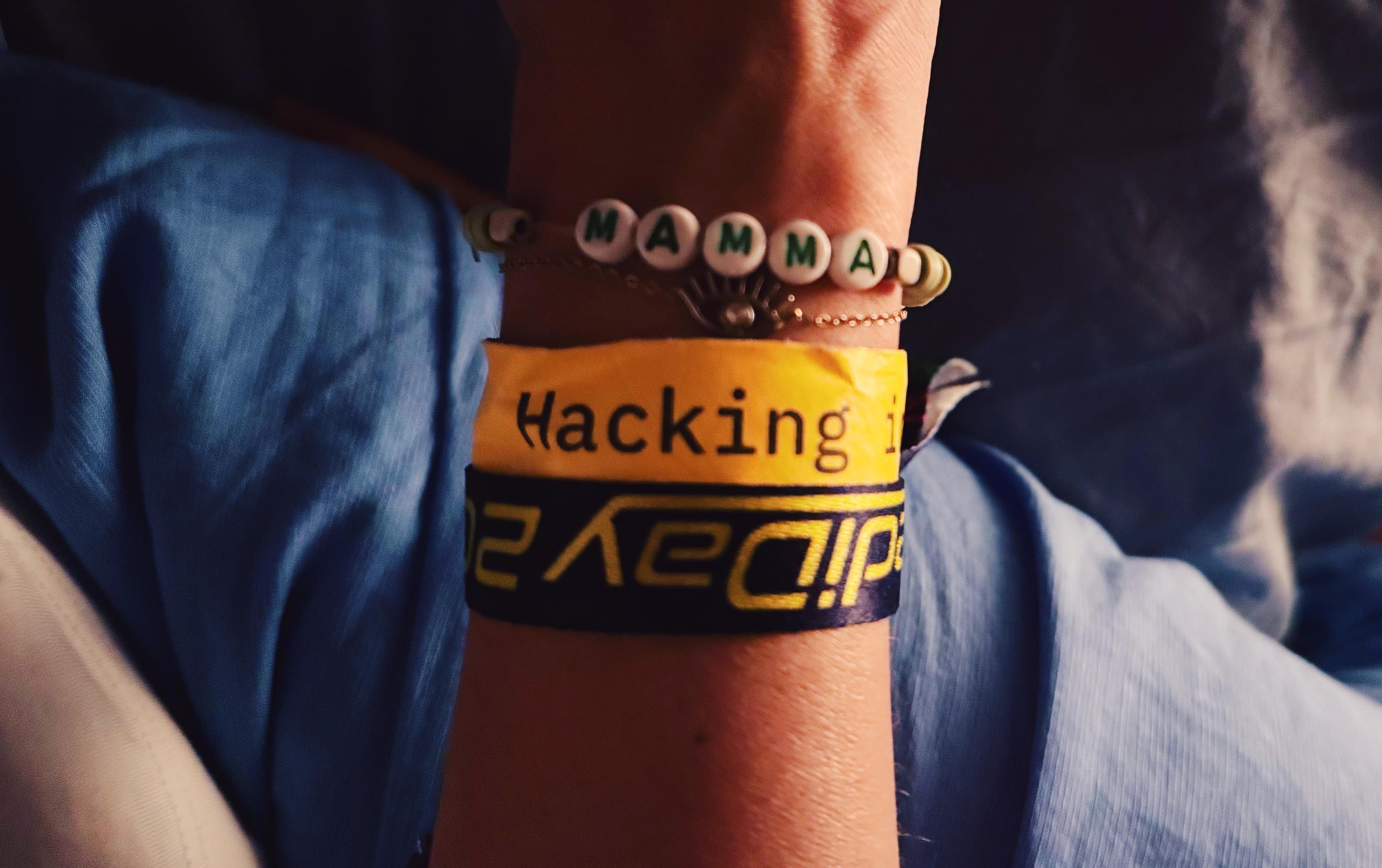 Picture of my wrist, seen from above. I’m wearing multiple bracelets: a homemade one that says “mamma” and below an eye bracelet, a yellow paper bracelet from Berlin Fedi Day that reads “Hacking” and below a purple bracelet (fabric) that reads « Fedi Day 2026 »