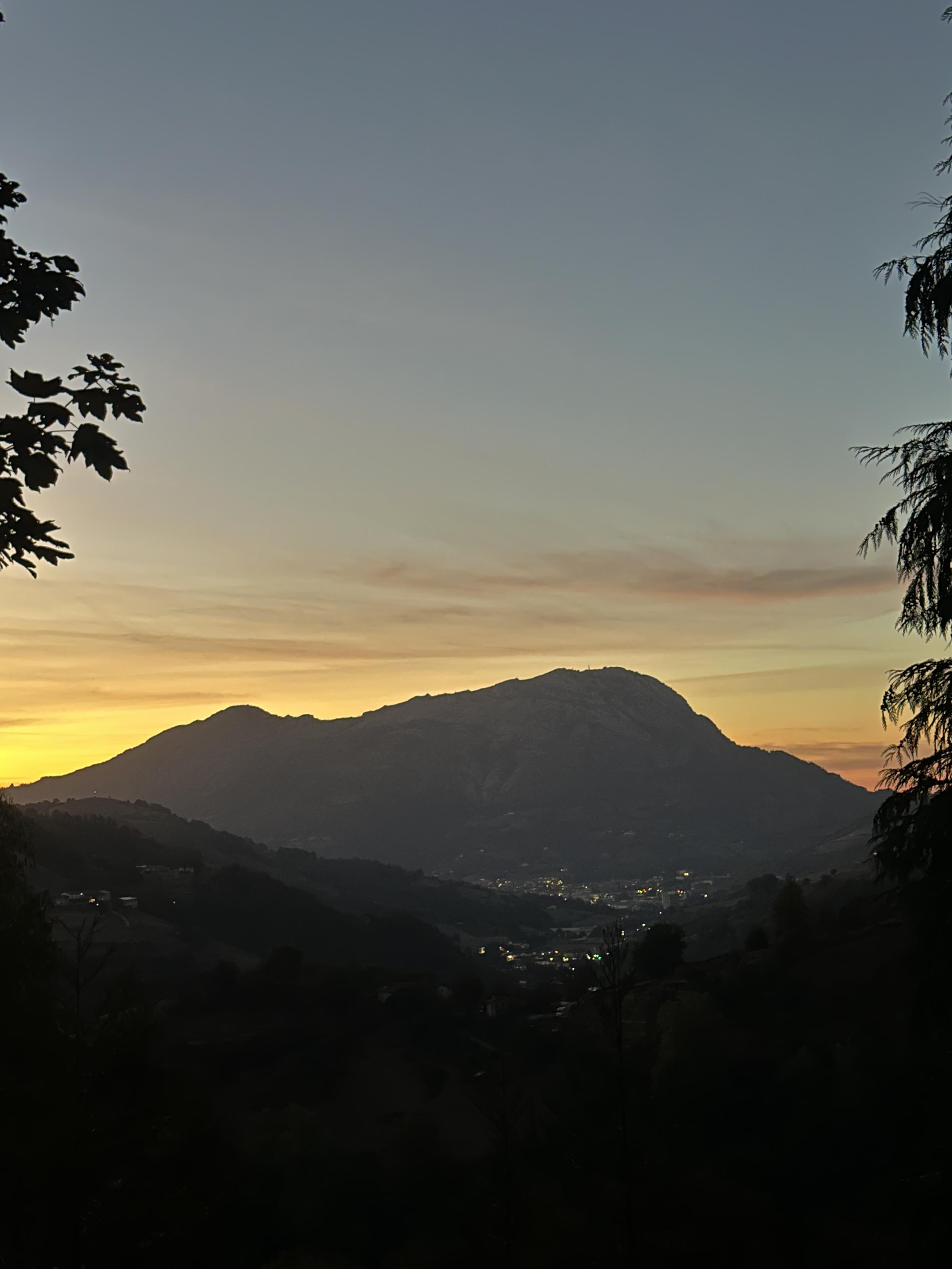 “Una foto de paisaje vertical y serena que captura una puesta de sol en las montañas. En el primer plano, siluetas oscuras y nítidas de ramas de árboles enmarcan los lados superior izquierdo y derecho de la composición, creando profundidad. En el centro, una gran montaña con múltiples picos se destaca como una silueta oscura y detallada contra el cielo de la puesta de sol.
El cielo es un degradado sutil y suave que comienza con un amarillo vibrante y cálido en el horizonte, justo detrás de la montaña, y se desvanece gradualmente a través de tonos melocotón y rosa suave antes de fundirse en un azul pálido y frío en la parte superior. Hay unas pocas nubes delgadas y rosadas esparcidas por el cielo medio.
En el valle oscuro y sombrío situado por debajo de la montaña y entre las colinas del primer plano, se aprecian las luces de un pequeño pueblo, parpadeando como diminutos puntos amarillos y blancos. La oscuridad de las colinas del primer plano contrasta fuertemente con la luz del cielo y
