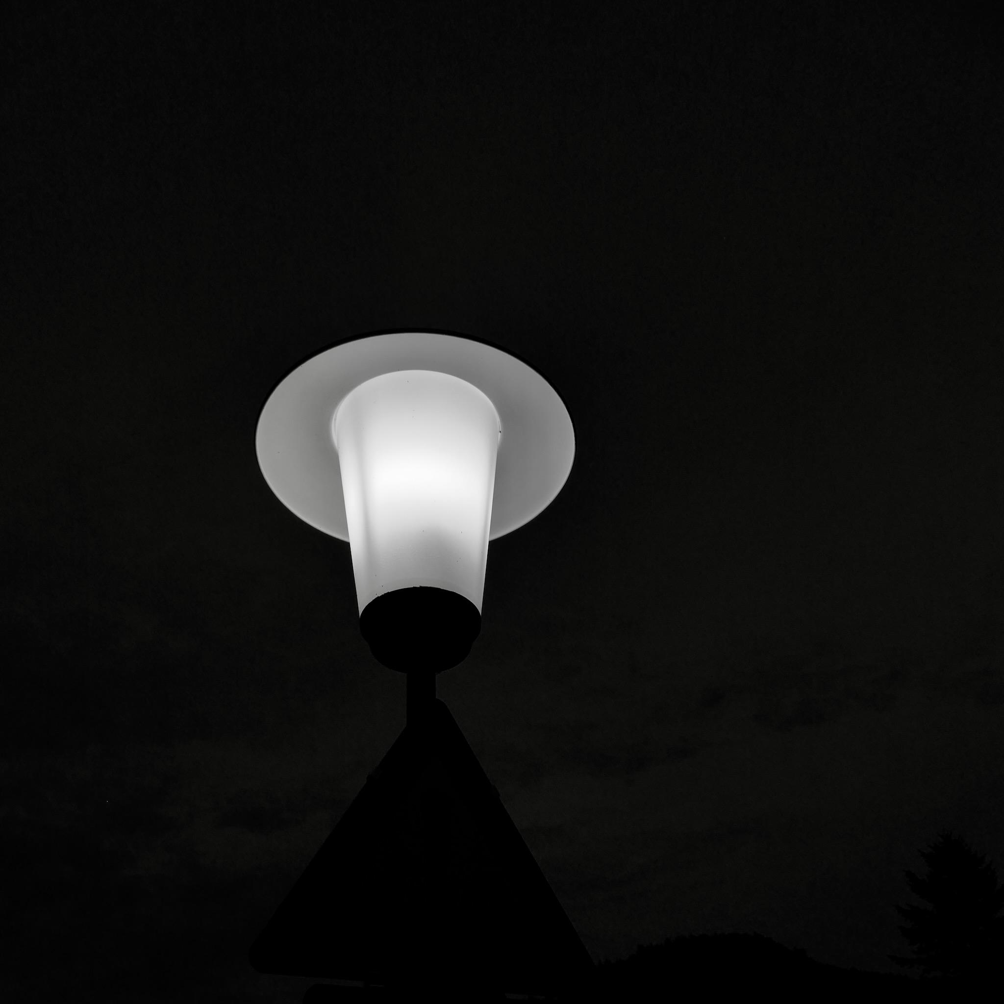 Photo of a streetlight in darkness. It is illuminated. The lower part is a cone of white light, up there is a round sheet of metal sitting like a hat. Otherwise the picture is black. Photo is black and white.