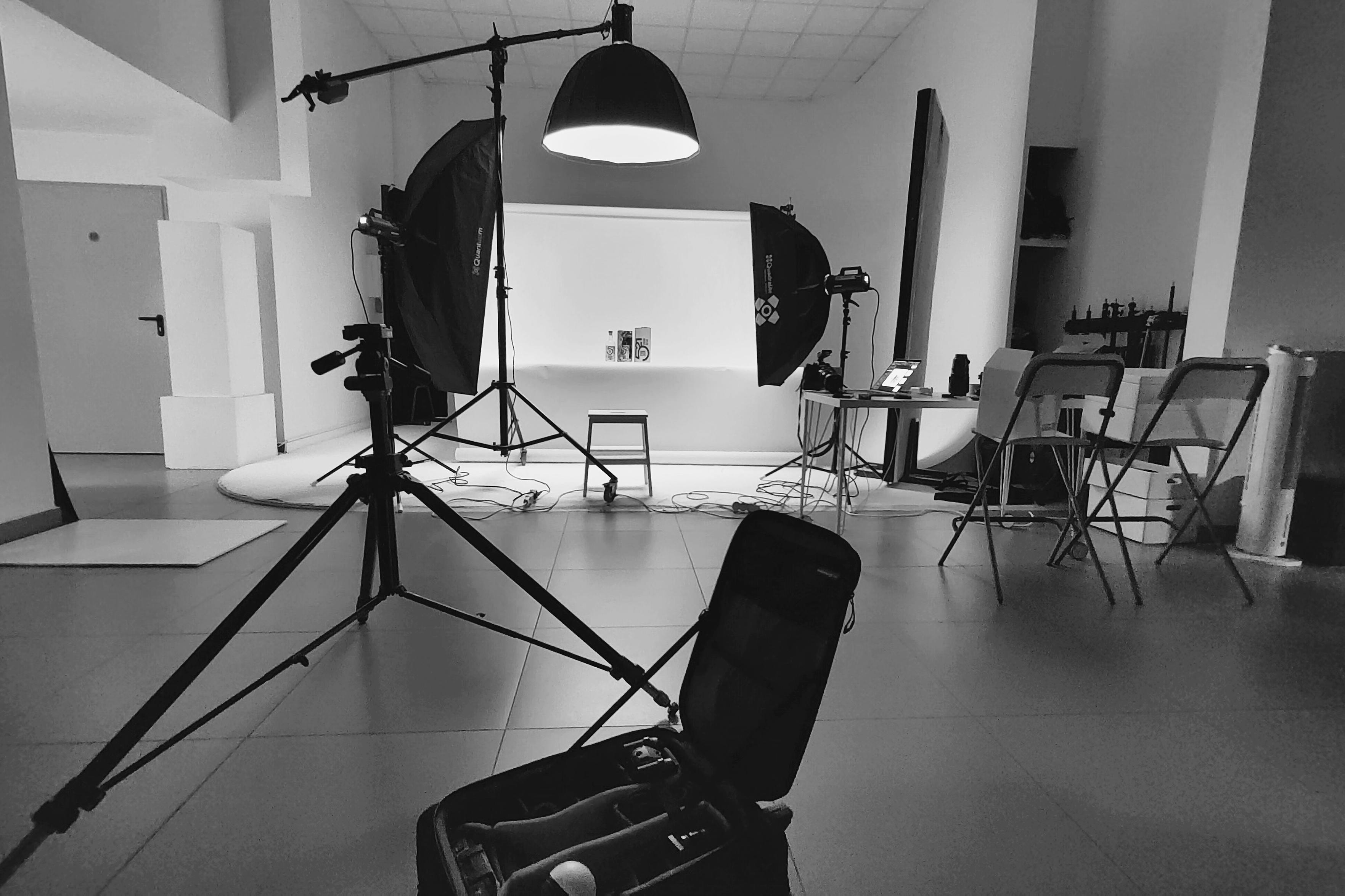 To zdjęcie przedstawia kulisy profesjonalnej sesji fotograficznej w studiu - Studio Stawki, wykonane w konwencji czarno-białej. W centralnym punkcie znajduje się tło z białej, wyprofilowanej ściany (cyklorama), która służy do fotografii produktowej. Nad tłem zamontowana jest duża lampa z czaszą oświetleniową. Po lewej stronie tła stoi duży softbox z gridem, a po prawej mniejszy softbox z parasolką dyfuzyjną. Przed tłem stoi mały metalowy stołek ze stojakiem na produkty, na którym leży mały przedmiot. Po lewej stronie kadru, na pierwszym planie, widać duży statyw fotograficzny i otwartą torbę na sprzęt fotograficzny, która jest lekko rozmyta. Po prawej stronie, w głębi, stoi stół z laptopem, aparatem fotograficznym i trzema składanymi krzesłami. Cała scena jest oświetlona lampami studyjnymi, a na podłodze widać kable i sprzęt. Całość tworzy profesjonalny klimat.