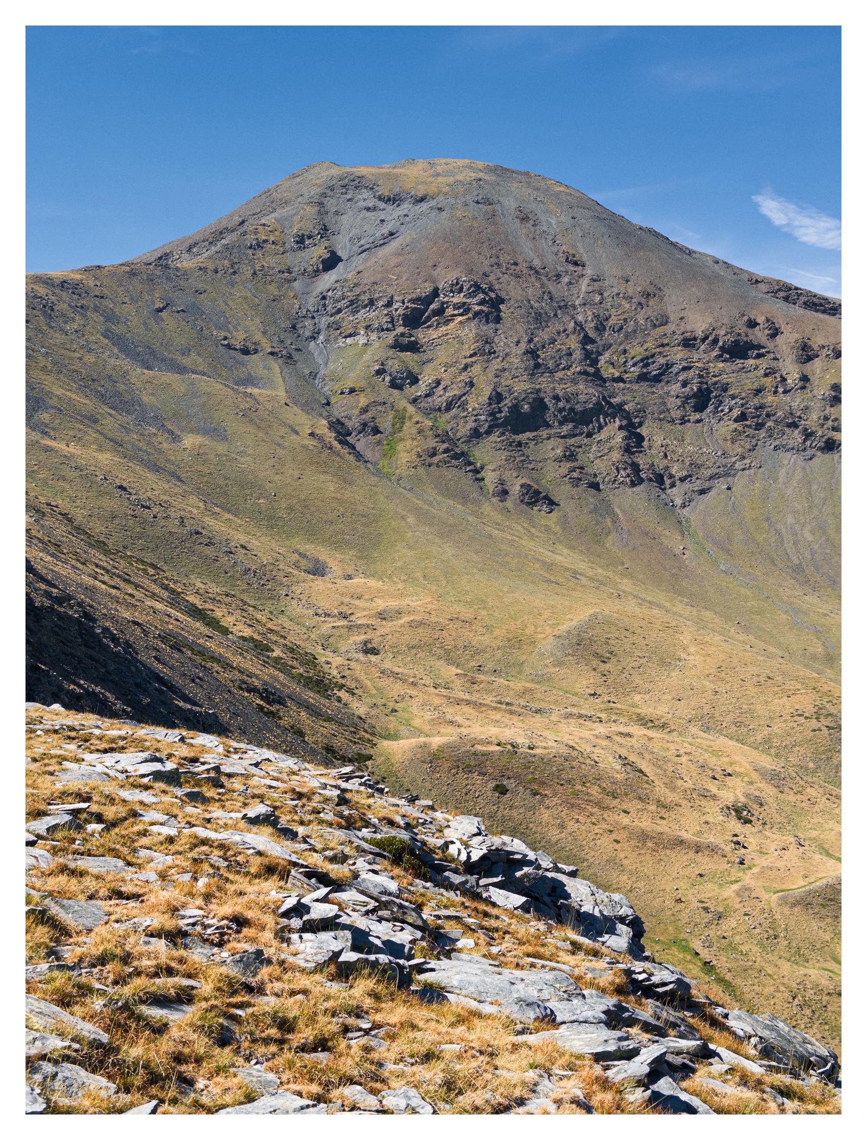 A colour photograph of an end-of-summer mountain landscape under a clear blue sky. A broad, rounded rocky mountain summit with vertical rock formations and erosion channels stands in the background occupying the upper half of the frame. In the foreground, a steep sloped terrain covered with fragmented pale flat rocks, probably schists, and dry grass. In between, a valley with undulating slopes covered by a few patches of green grass intertwined with a majority of golden dry alpine vegetation.