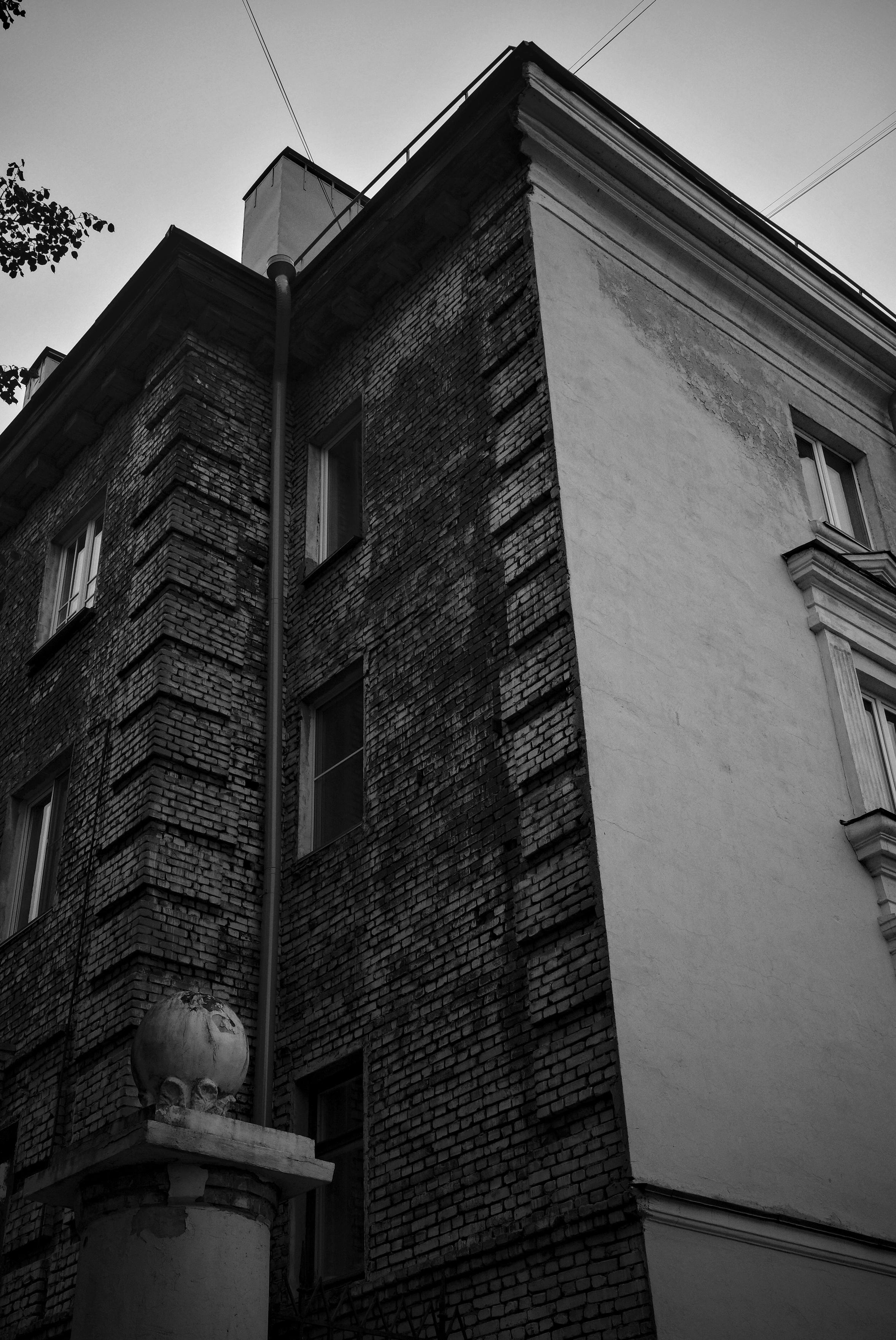 This black-and-white photograph captures a fragment of an old city building, shot from a low angle looking upward. The frame is built on the contrast of two textures: on the left and in the center — dark, rough brickwork with deep joints and traces of time; on the right — a smooth, light plastered wall. A vertical drainpipe divides the composition and guides the viewer's eye. Windows with white frames are visible on the facade. In the lower left corner, in the foreground, a concrete pillar with a broken sphere stands out — a detail that adds a feeling of abandonment to the frame. Above, against the gray sky, thin wires and the edge of the roof are visible. The image conveys the atmosphere of an old city, where architecture preserves traces of time.