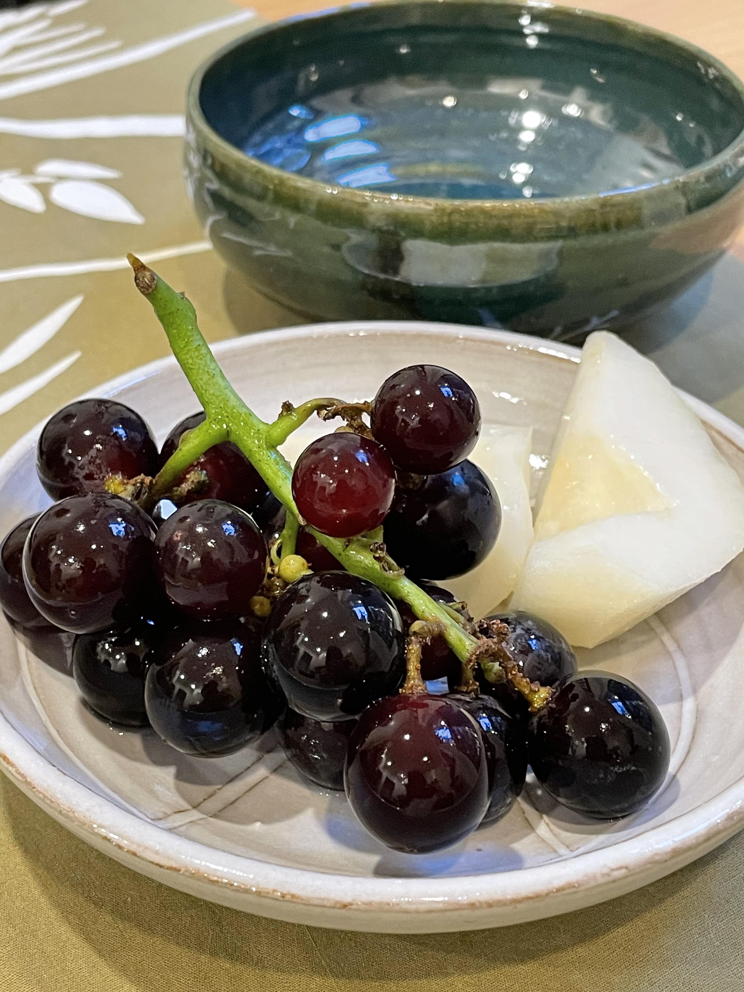 A bunch of grapes and peeled, quartered pear on a white plate, with a green bowl behind it on a table