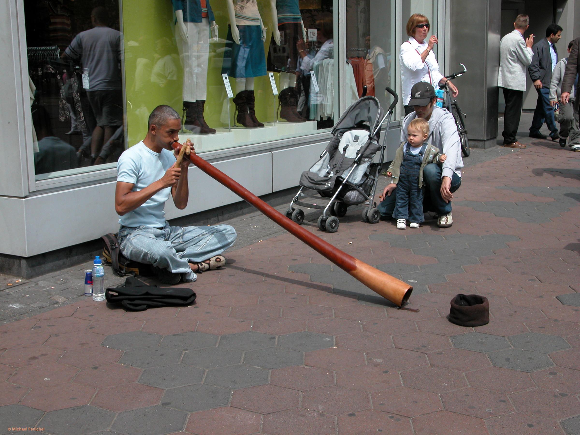 Photograph by Fenichel

Sitting on a wide sidewalk with tiled patterns, a man in white shirt and jeans sits cross-legged and plays a didgeridoo, his back to a store window which shows reflections of the street as well as the store display To the right, watching with curiosity, is a young boy, stroller nerby and his Dad, crouching behind the boy.
