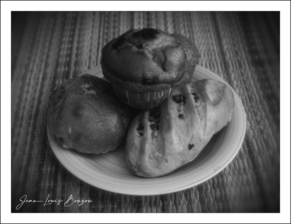 The image is a black and white, close-up shot of three baked goods arranged on a white plate, which is placed on a textured surface. The baked goods appear to be a muffin, a small bread roll, and a small loaf or bun.
The muffin is positioned at the top and center of the plate, dominating the composition. It is dark in color, with visible creasing from its paper liner. The top of the muffin shows subtle texture and some darker specks, suggesting chocolate chips or dried fruit.
To the left of the muffin is a round, slightly irregular-shaped bread roll. It has a smooth surface with some lighter areas and subtle indentations, indicating it might be slightly flattened.
To the right and slightly forward of the muffin is a small, elongated loaf or bun. It has a lighter crust with a few visible darker inclusions, possibly raisins or chocolate chips, embedded in the dough. A series of parallel score marks are visible on its surface, characteristic of bread making.
The plate is white with subtle
