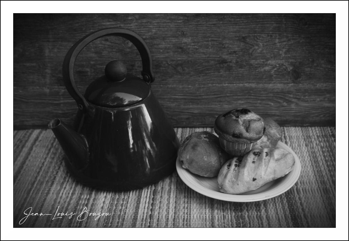 The image is a black and white still life featuring a tea kettle and a plate of baked goods set against a rustic wooden background.

The main elements are a large, dark, possibly red, tea kettle positioned on the left side of the frame. It has a curved handle and a prominent spout. To the right of the kettle, on a white plate, are several baked items. These include what appears to be a muffin in the foreground, with a dark, slightly burnt top, and several other round buns or small loaves of bread. One of the bread items in the front has visible score marks. The baked goods are arranged in a cluster on the plate.

The scene is set on a textured surface, likely a placemat or woven cloth, with a distinct striped pattern. Behind the objects is a dark wooden backdrop, with visible grain and planks, adding to the rustic feel of the image.

The color palette is monochromatic, relying entirely on shades of gray, black, and white. The black and white treatment emphasizes the textures and shapes