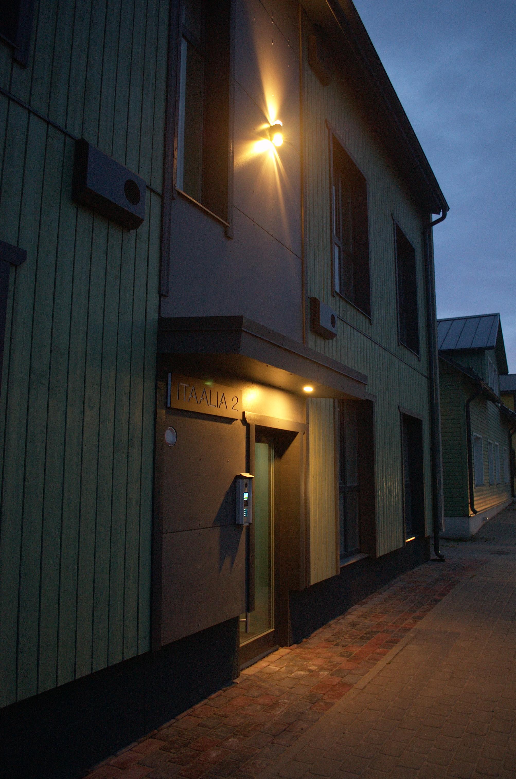 Photo of the facade of a pale green house, taken at an oblique angle. A light between the second storey windows casts fans of yellow light on the wall. A smaller light under an awning lights up a glass door. The sidewalk in front of the house is covered in grey paving stones, but a narrow strip along the wall is made of reddish cobbles.