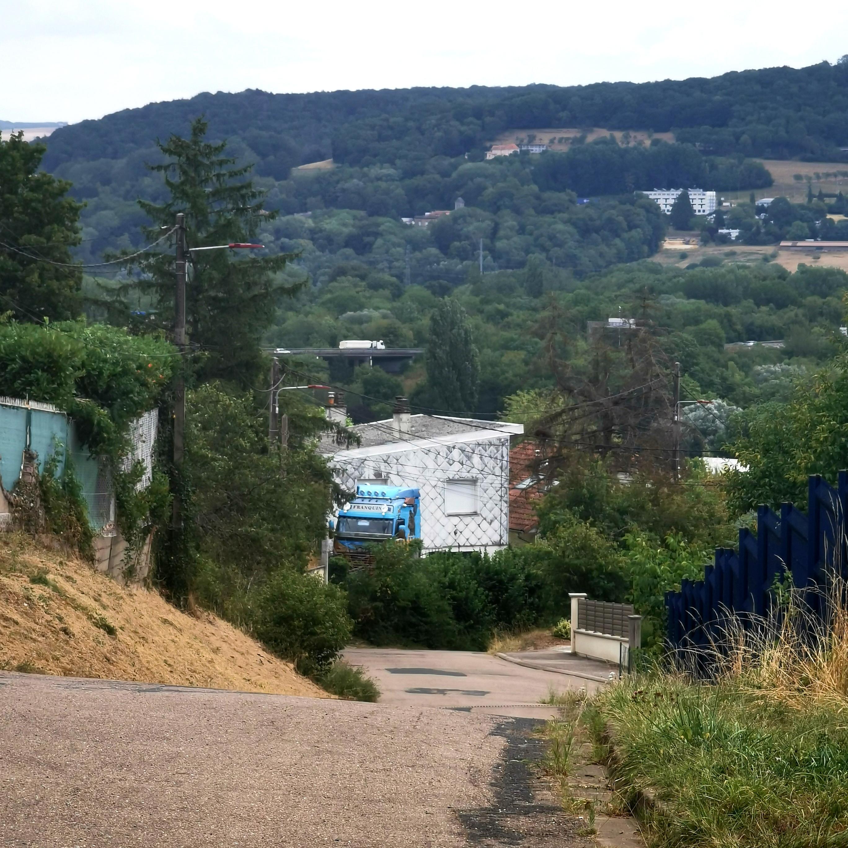 Vallée sous une forte colline, habitat épars, au premier plan une maison devant laquelle est garé un camion.