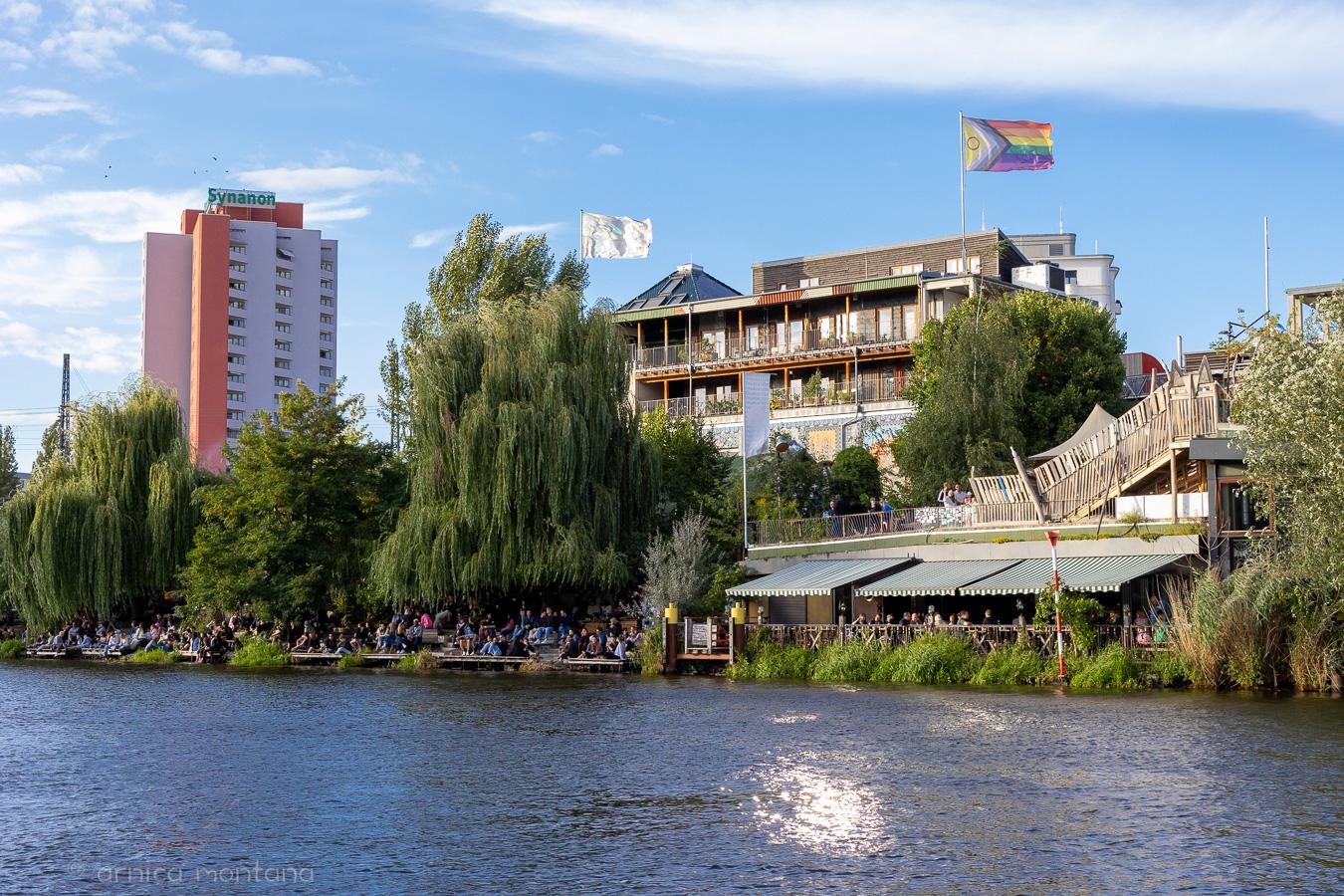 The photo shows a restaurant on the banks of the Spree in Berlin, many people are sitting in the sun and a Pride flag is flying above them all. Green trees line the riverbank, the sky is blue and a high-rise building can be seen on the left.