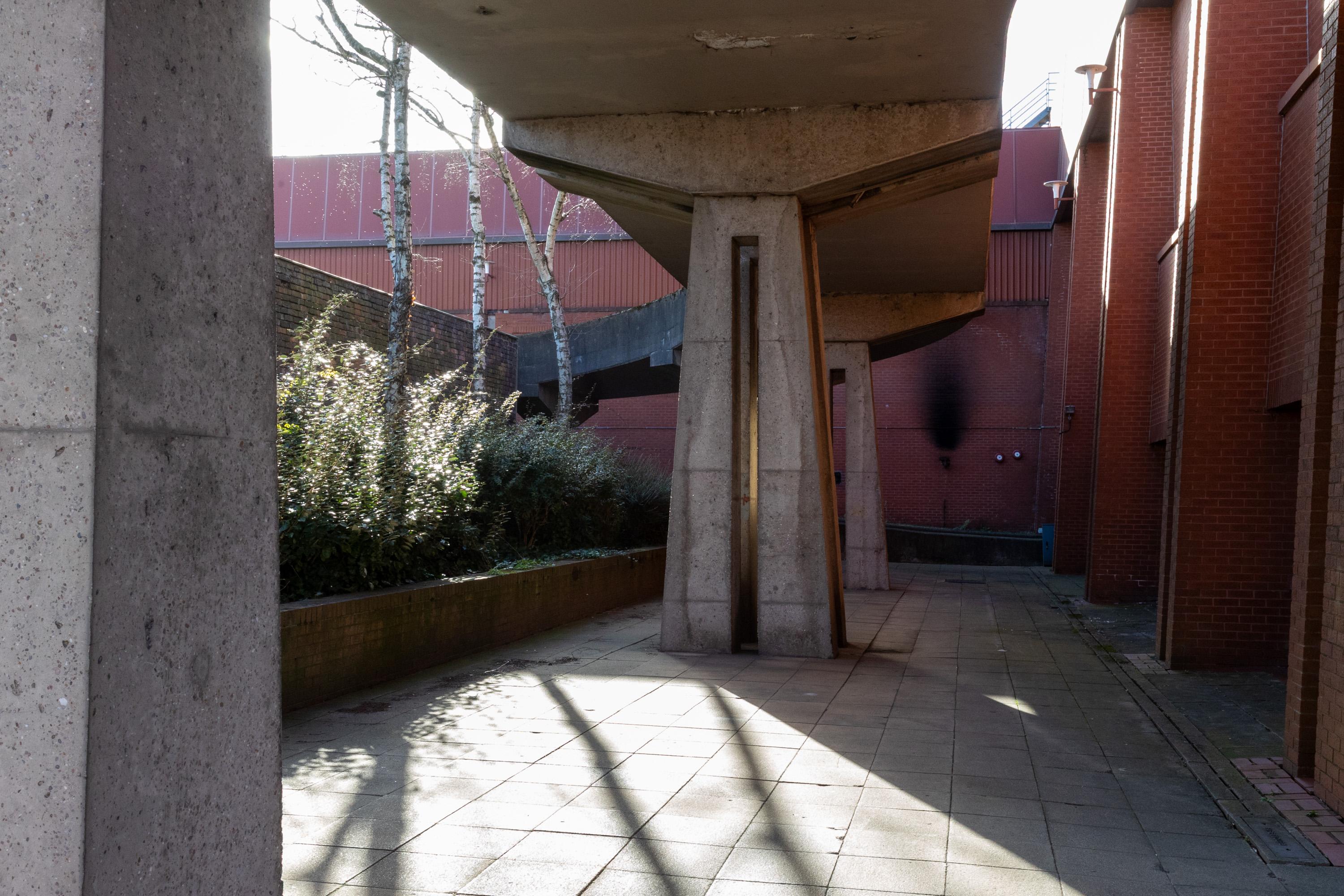 A concrete walkway with thick pillars and overhead structure casts shadows on the paved path below. Brick buildings and greenery line the left side, whilst sunlight streams in, highlighting the geometric architectural shapes.