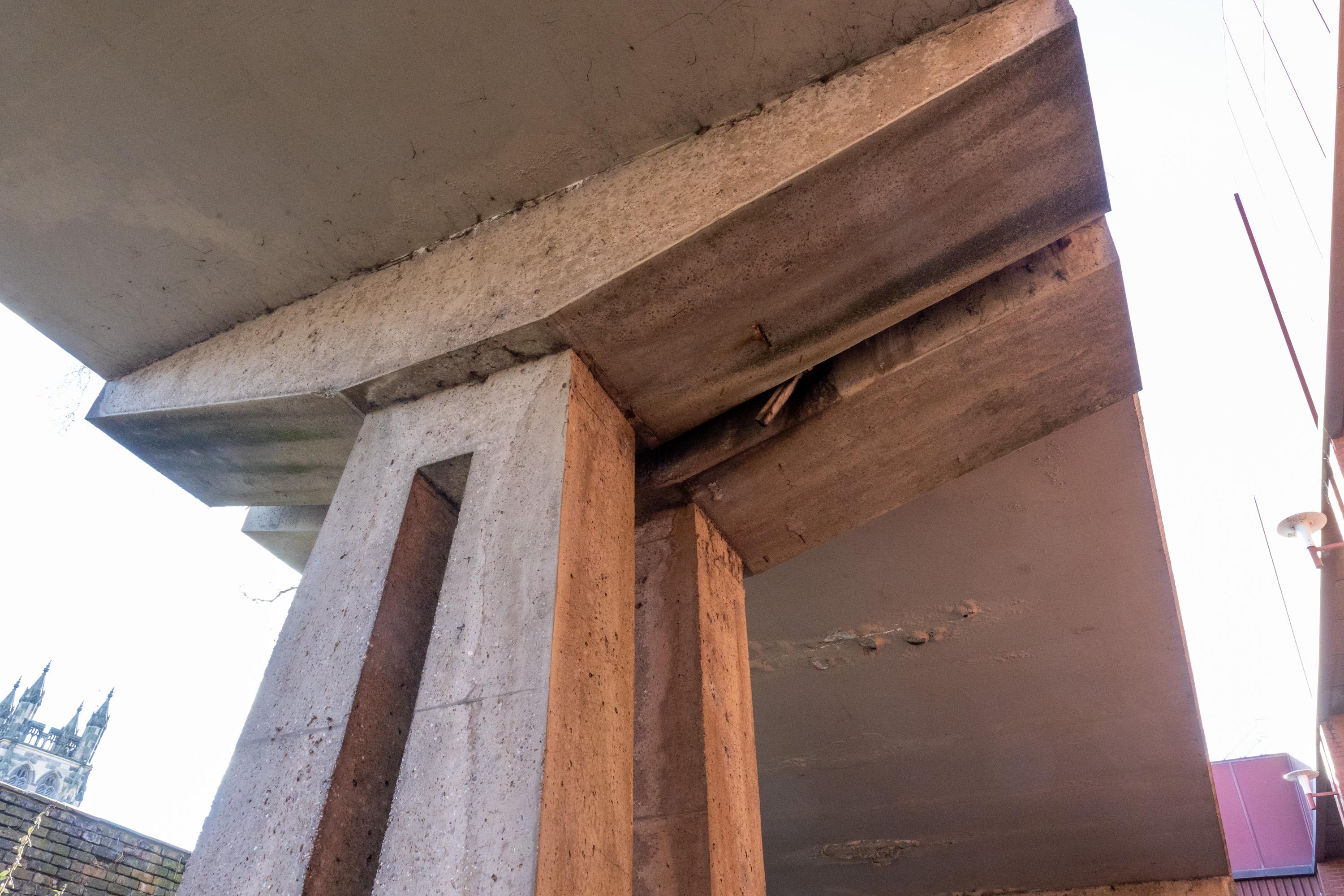 Close-up view from below of a large concrete overhang supported by a pillar. There are visible cracks, stains, and spalling on the underside and edges, suggesting structural wear and damage. The sky and part of a building are seen in the background.