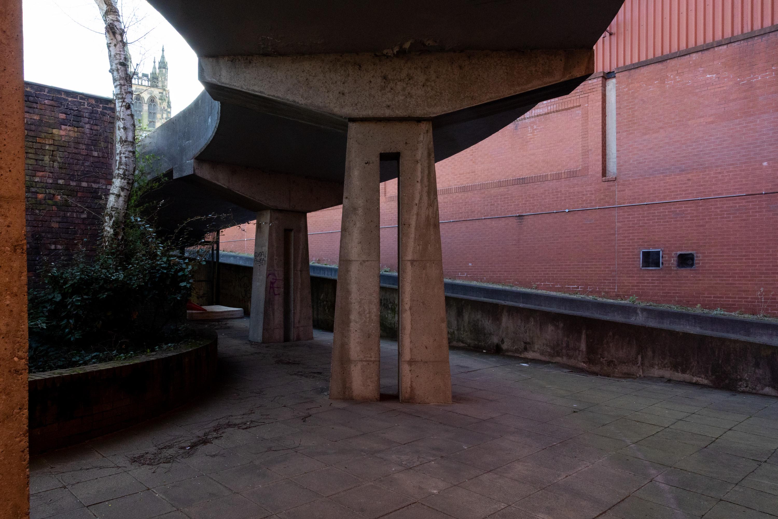 Concrete walkway with angular pillars underneath, casting shadows on paved ground. Brick walls on both sides; a round planter with a tree on the left. In the background, a church tower is partially visible above the wall. The scene appears empty and quiet.