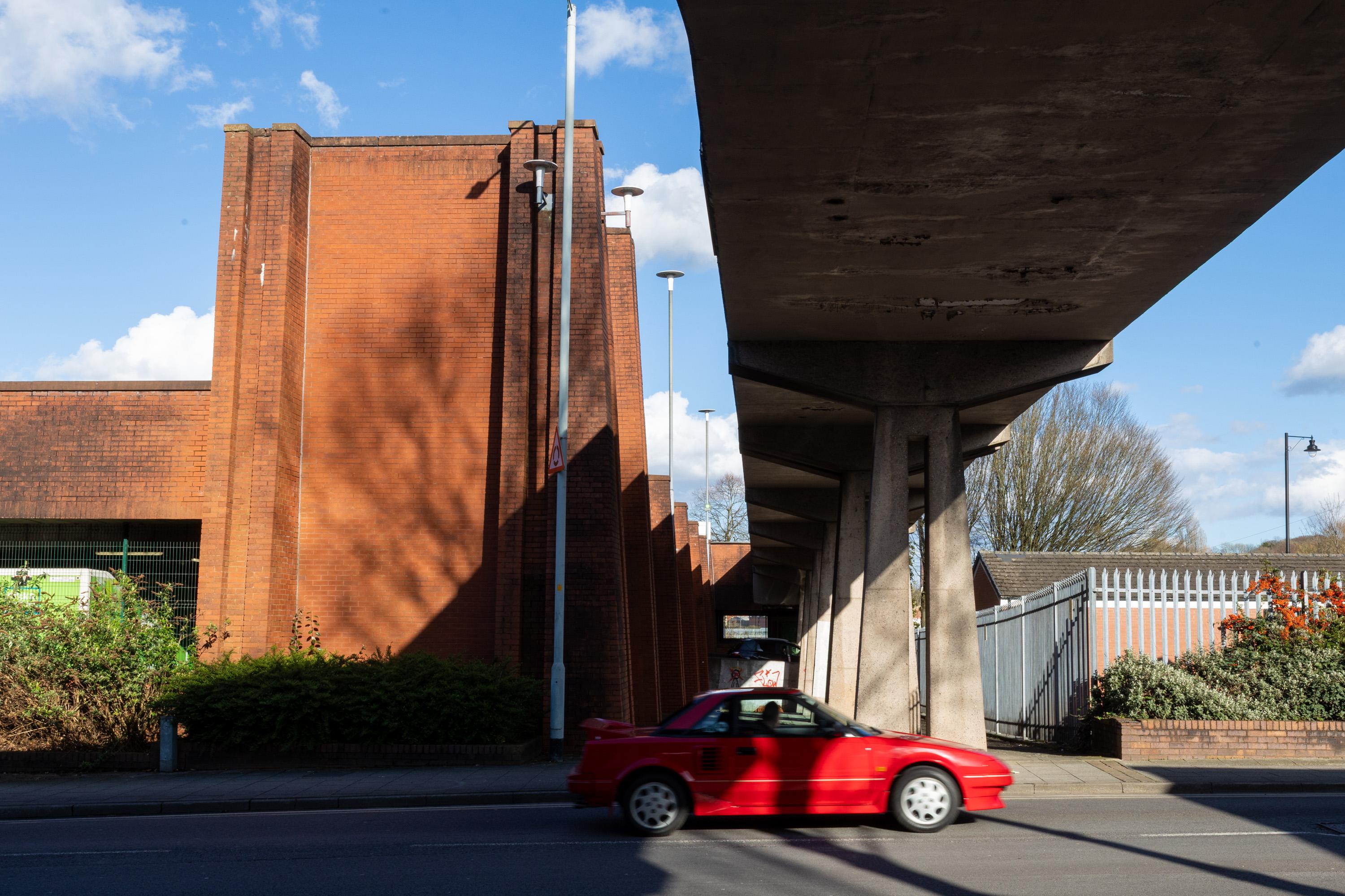 A small red convertible drives on a city street under a concrete flyover. Tall brick buildings cast shadows. Trees and bushes border the area. The sky is blue with scattered clouds. The scene is brightly lit by sunlight.