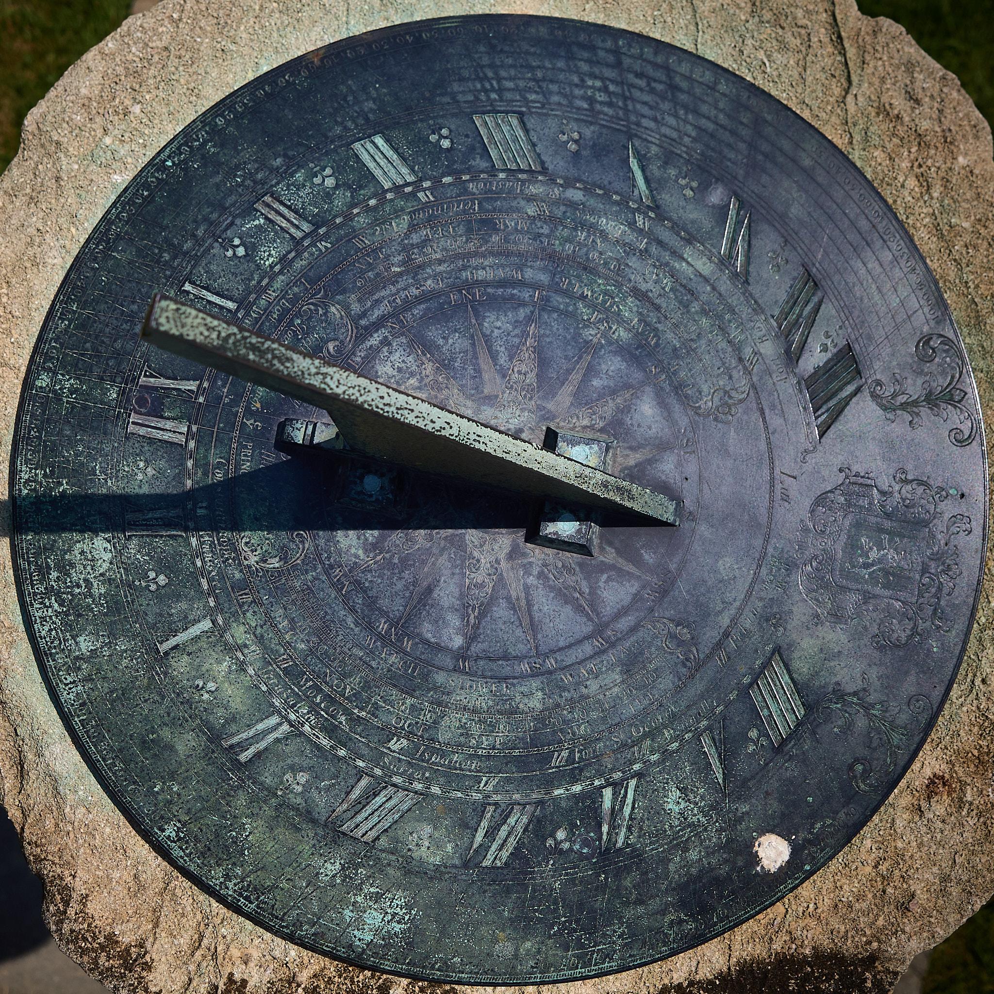 A sundial made of bronze is positioned on a stone base. The sundial features Roman numerals and decorative engravings. A shadow is cast by the gnomon, indicating the time.